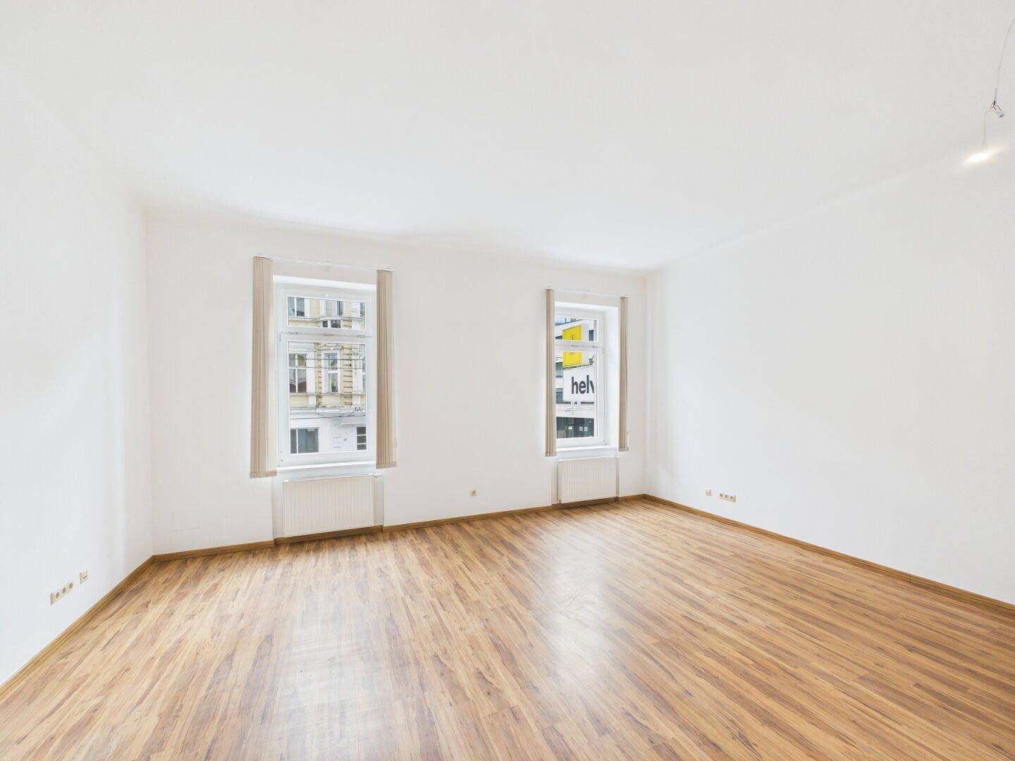 Helles Wohnzimmer mit Laminatboden und zwei Fenstern, bereit zur individuellen Einrichtung.