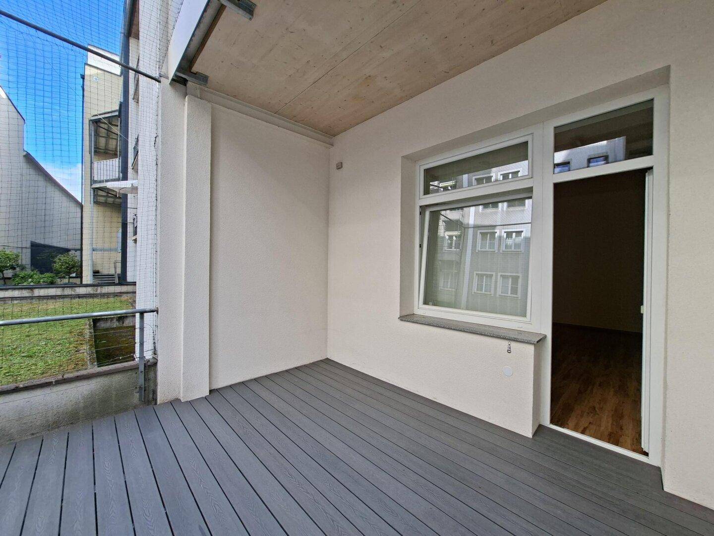 Heller Balkon mit grauen Dielen und Blick auf den begrünten Innenhof.