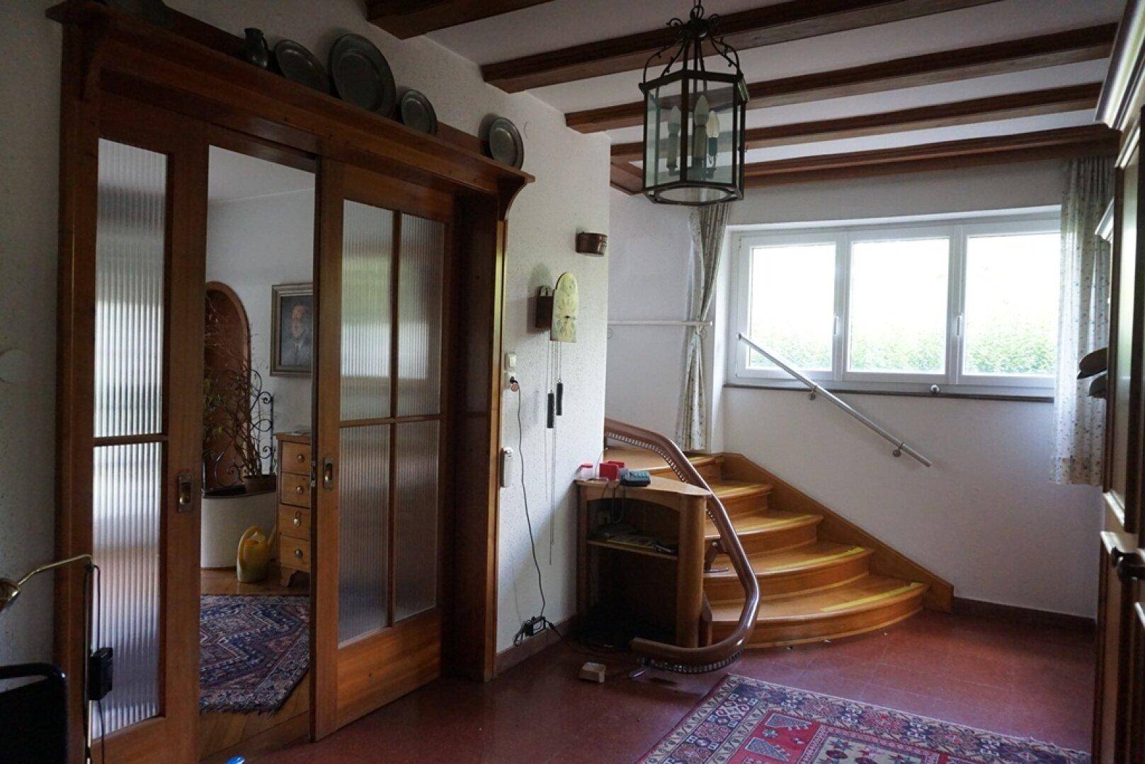 Heller Flur mit Holztreppe, großen Fenstern und rustikalen Deckenbalken.