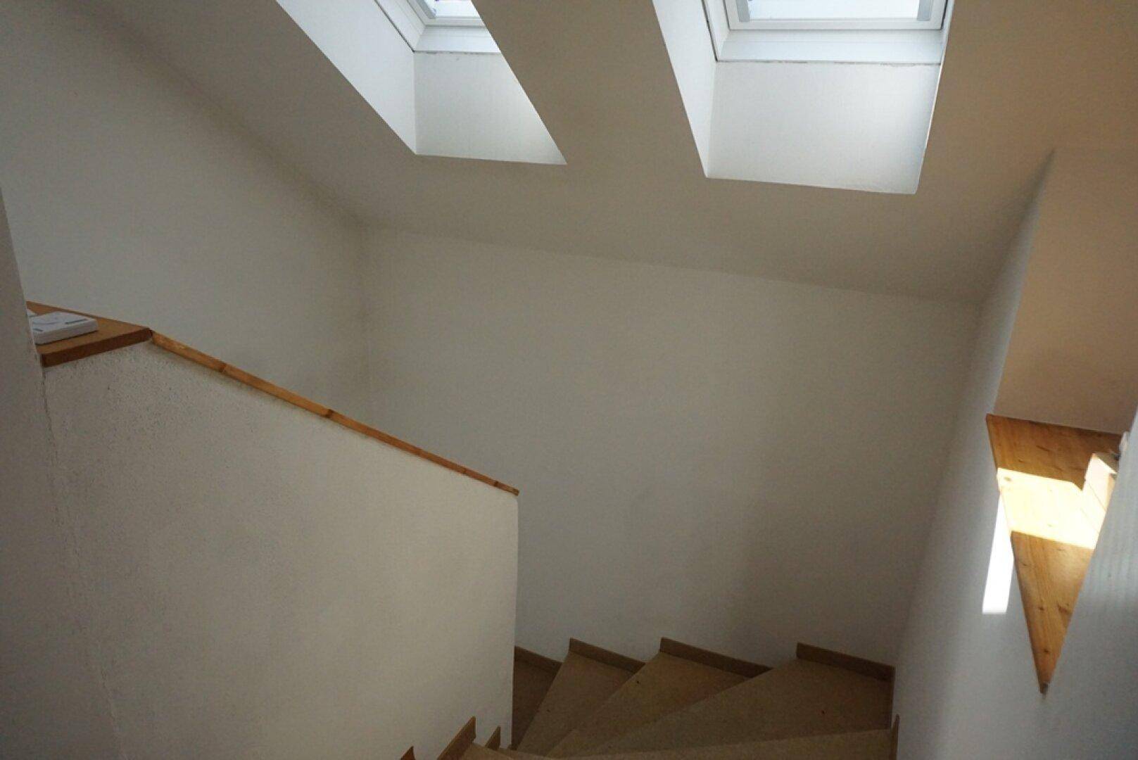 Treppenhaus mit Holzgeländer und zwei Dachfenstern, die für viel natürliches Tageslicht sorgen.