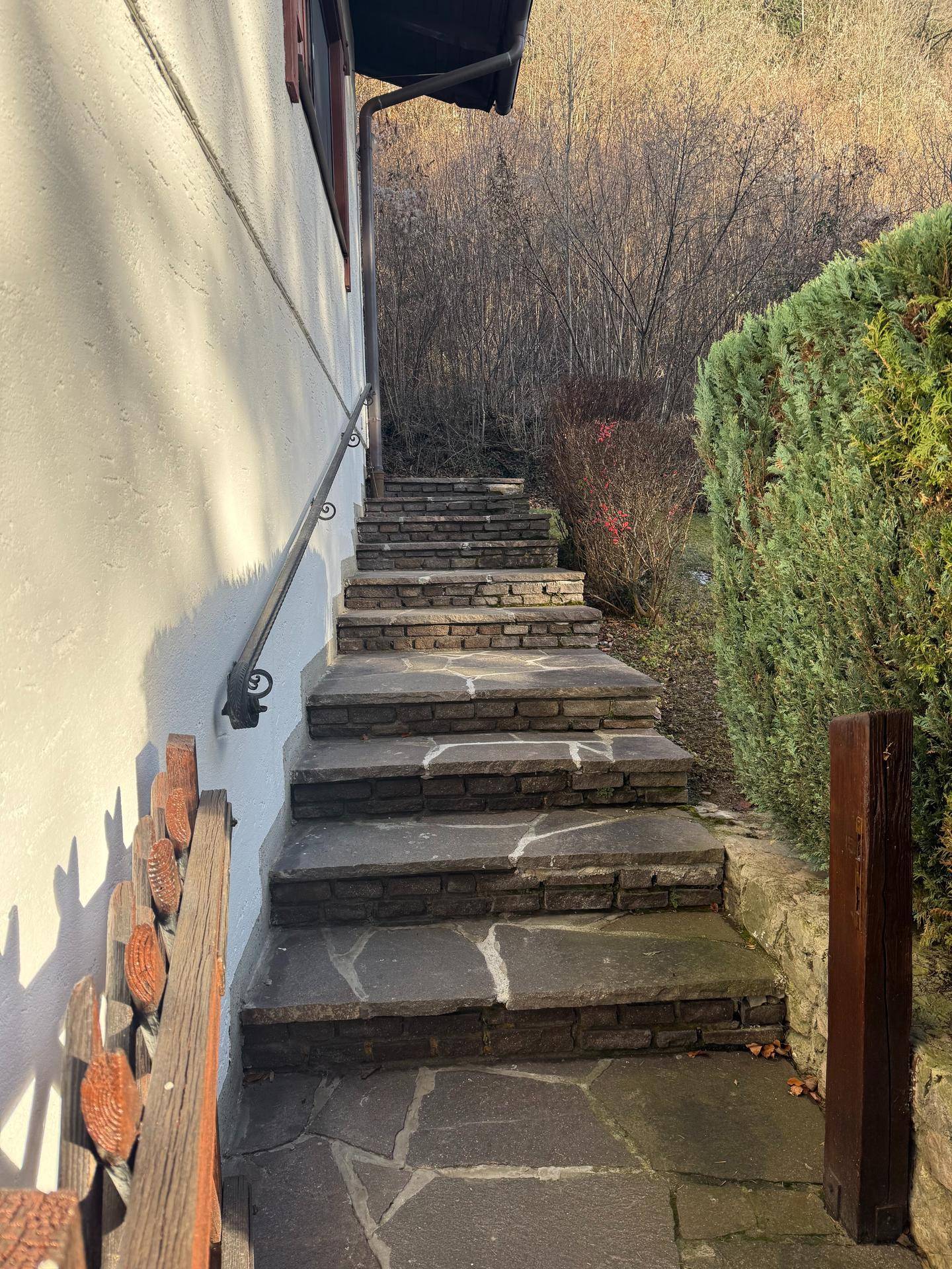 Steintreppe mit Handlauf entlang der Hauswand, umgeben von Hecken und Bäumen.