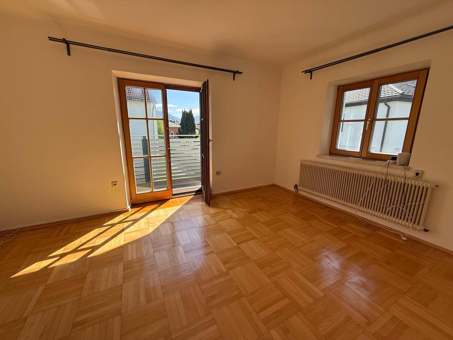 Lichtdurchflutetes Zimmer mit klassischem Parkettboden, weißen Wänden und Zugang zu einem Balkon.