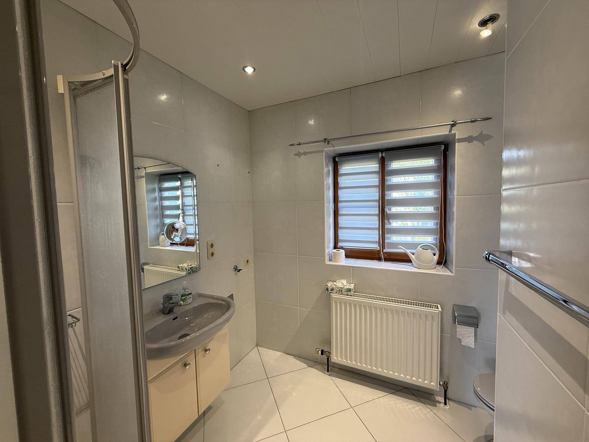 Gefliestes Badezimmer mit Duschkabine, Einzelwaschbecken, Heizkörper und Fenster mit Jalousien.