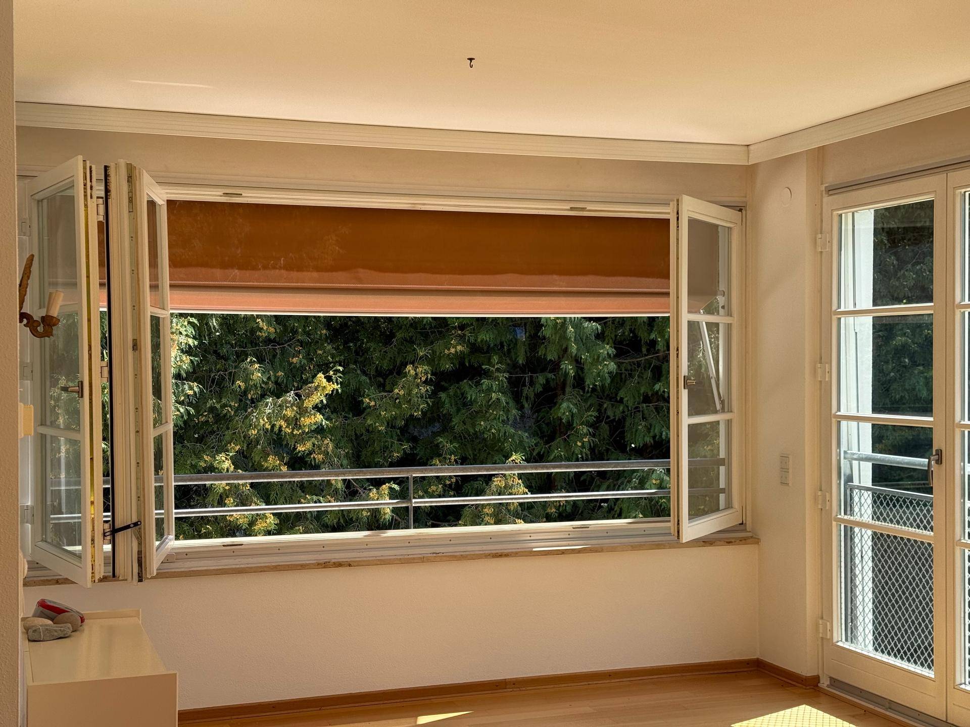 Heller Wohnraum mit Holzboden, großen Fenstern und direktem Zugang zu einem Balkon mit Grünblick.