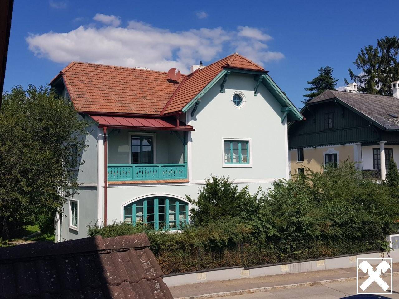 Seitenansicht der Villa mit Balkon, grünen Fensterrahmen und umgebender Vegetation.