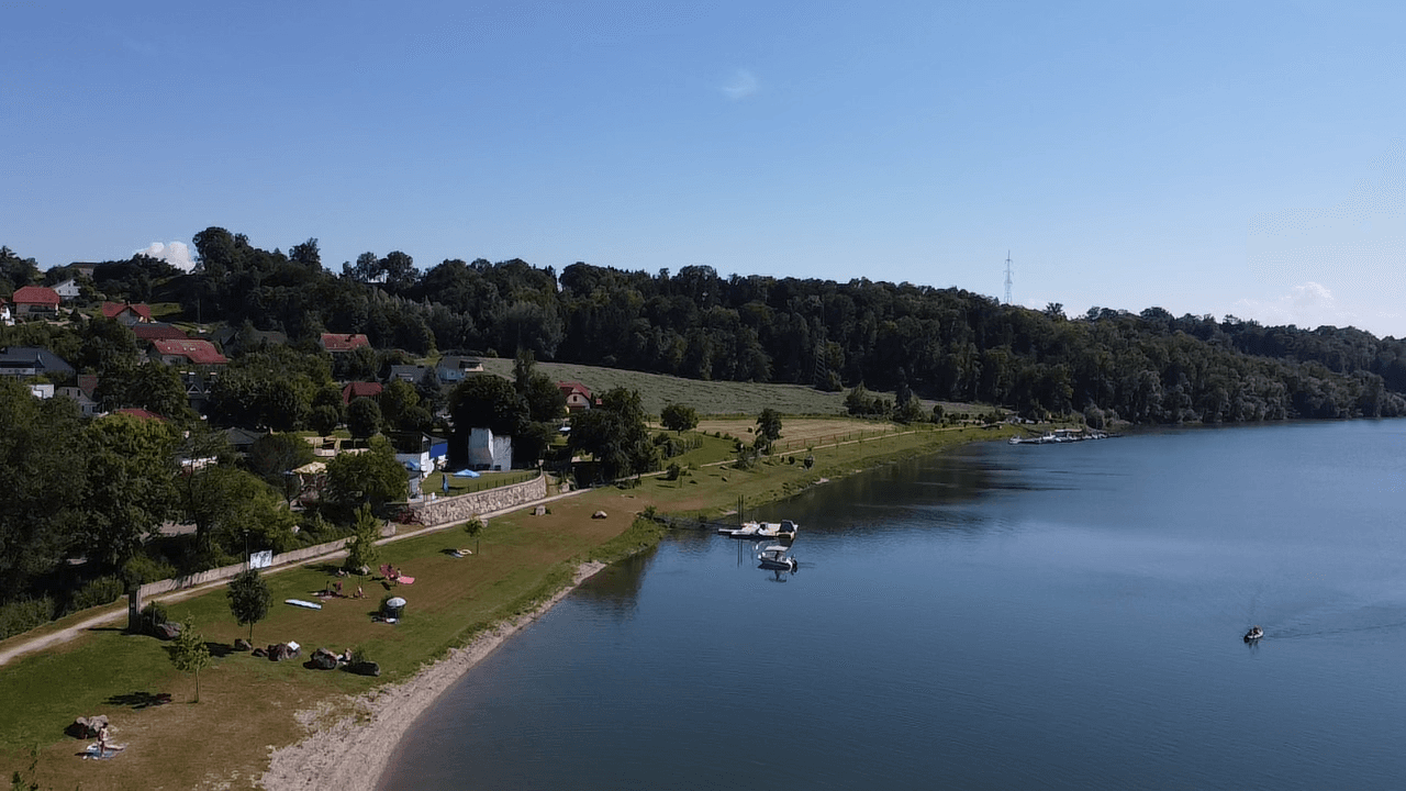Malerischer Seeblick mit Badestrand, bewaldeten Ufern und kleinen Booten am Horizont.