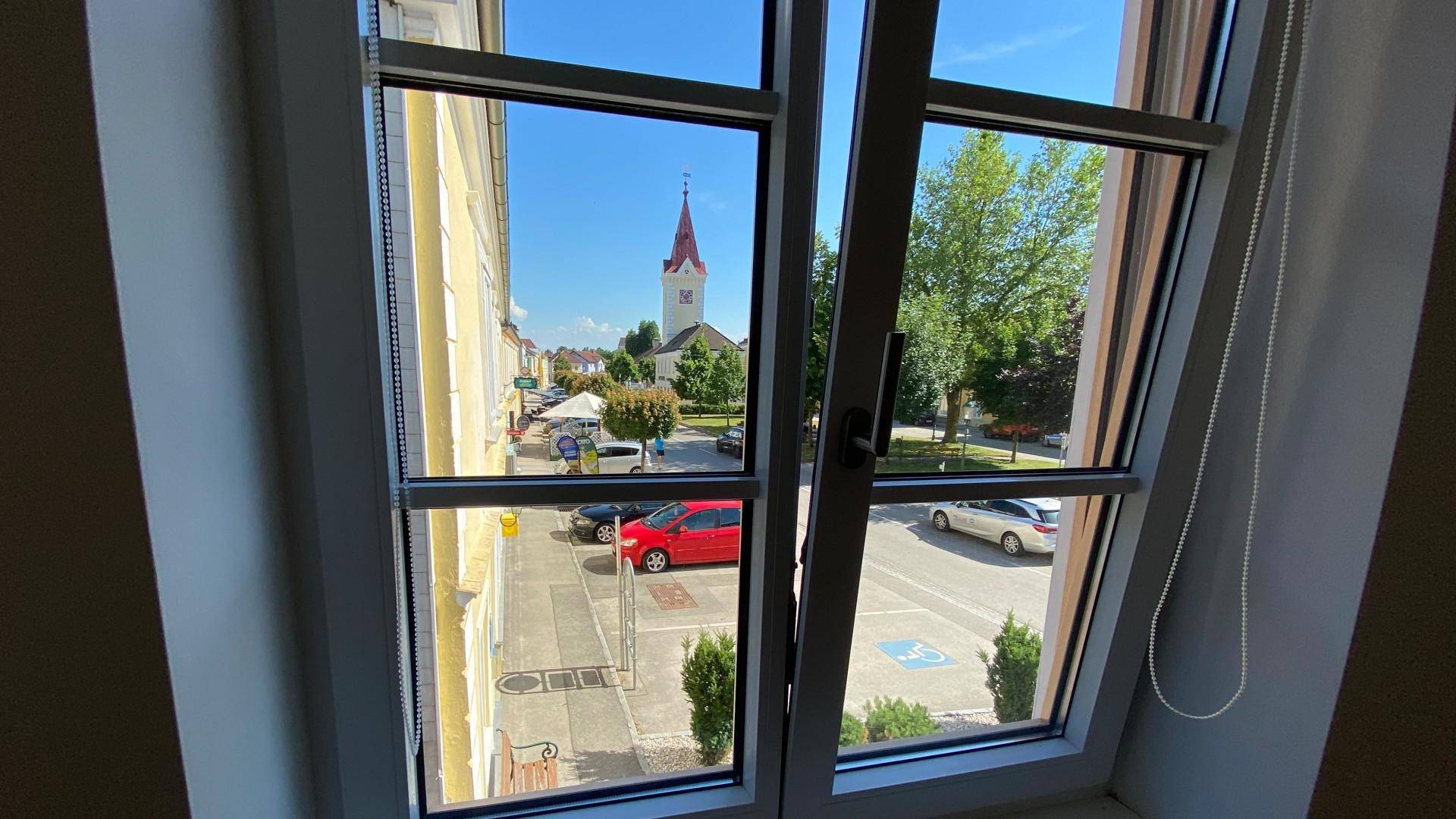 Fensterblick auf eine belebte Straße mit einem Kirchturm und grünen Bäumen im Hintergrund.