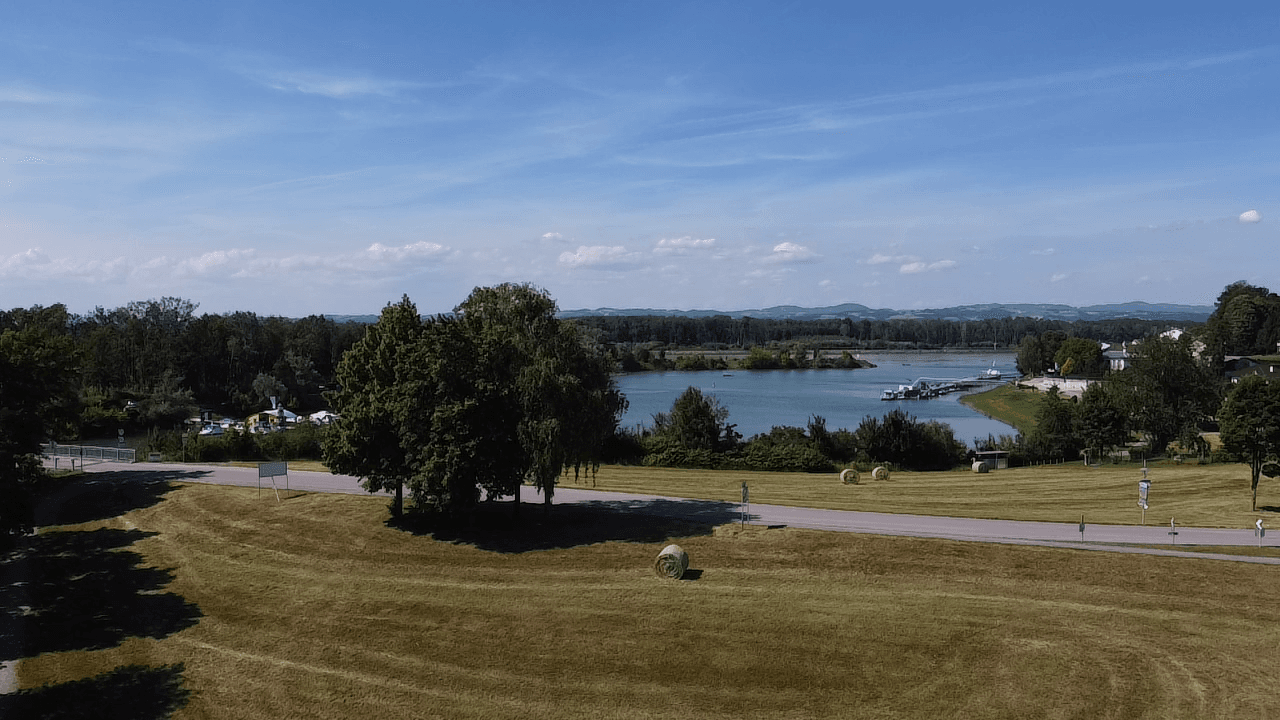 Weitläufiger Blick auf einen See mit grünen Wiesen, Bäumen und hügeliger Landschaft.