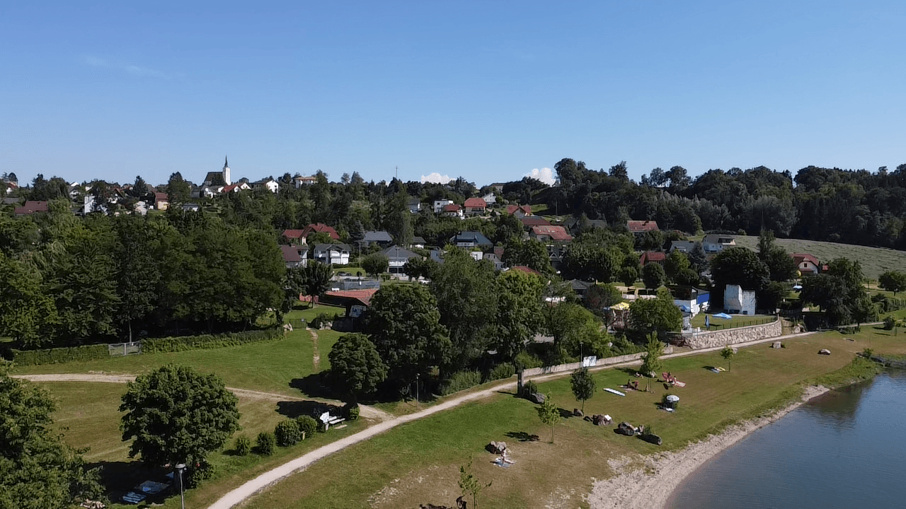 Panoramablick auf einen See mit Badestrand, bewaldeten Ufern und einem Dorf im Hintergrund.