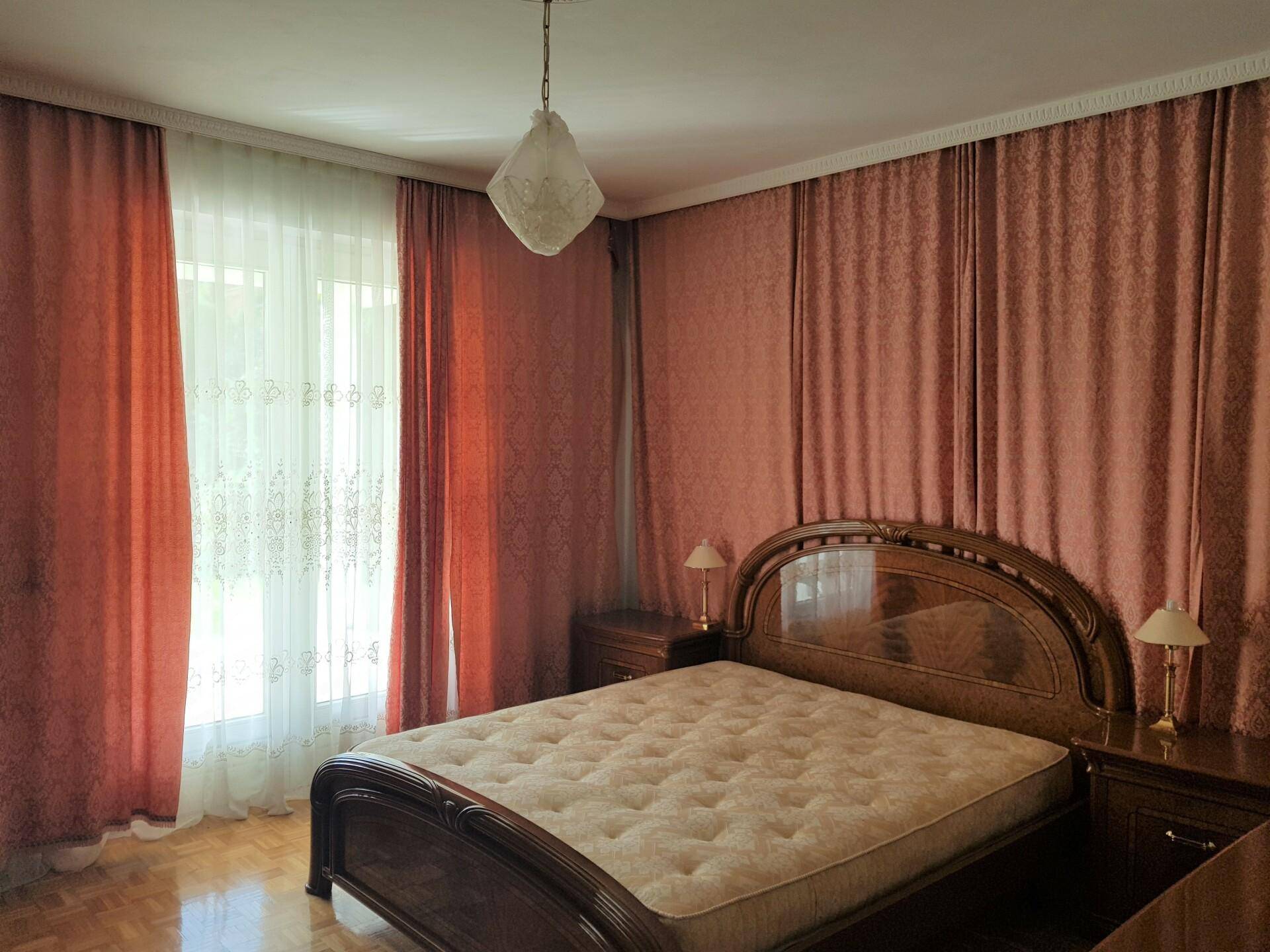 Helles Schlafzimmer mit klassischem Holzbett, Parkettboden und großen Fenstern mit Vorhängen.