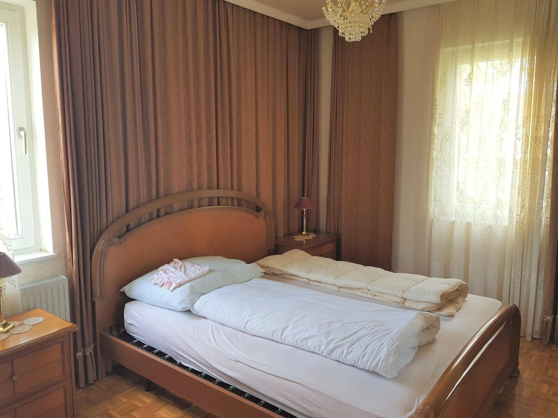 Helles Schlafzimmer mit klassischem Holzbett, Parkettboden und großen Fenstern mit Vorhängen.