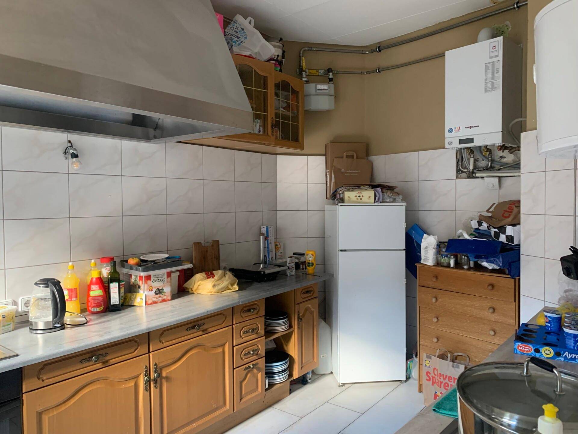 Funktionale Küche mit weißen Fliesen, Holzschränken, großem Dunstabzug und freistehendem Kühlschrank.