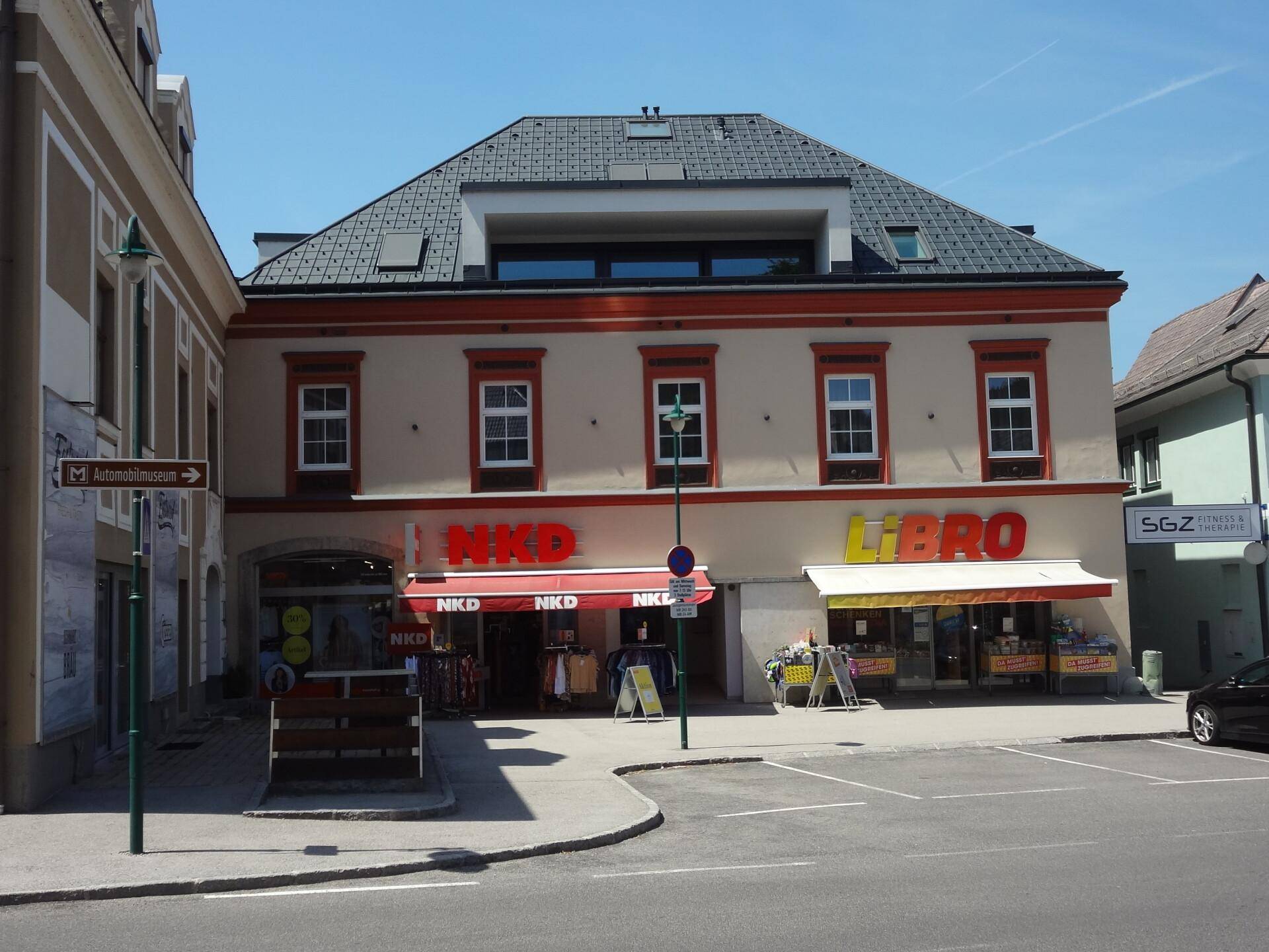 Weitläufige Straßenansicht eines Stadtplatzes mit Geschäften und gepflegten Blumenbeeten unter blauem Himmel.