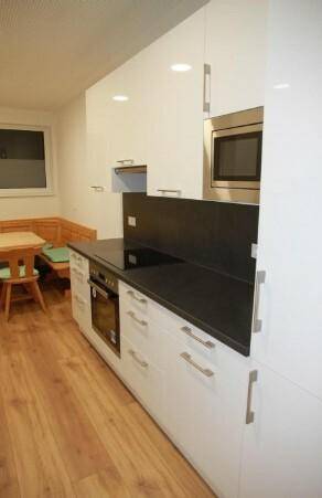Moderne Küche mit weißen Hochglanzschränken und dunklen Arbeitsplatten, angrenzend an einen kleinen Essbereich mit Fenster.