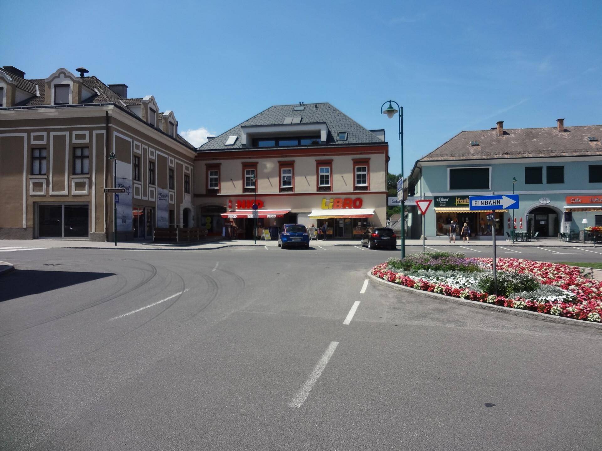 Weitläufige Straßenansicht eines Stadtplatzes mit Geschäften und gepflegten Blumenbeeten unter blauem Himmel.