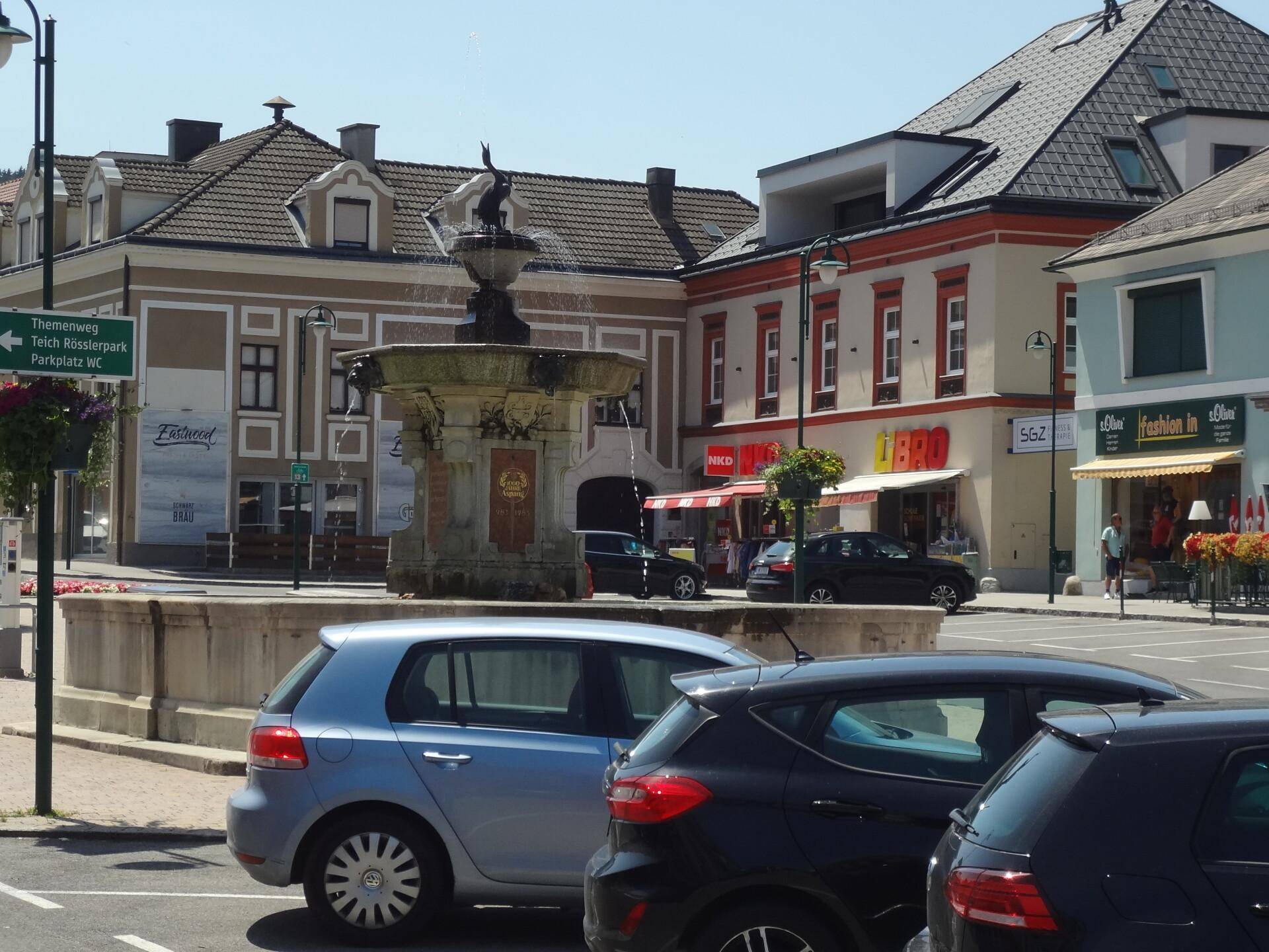 Panoramablick auf einen Stadtplatz mit historischen Gebäuden, einem Kirchturm und Blumenbeeten unter blauem Himmel.