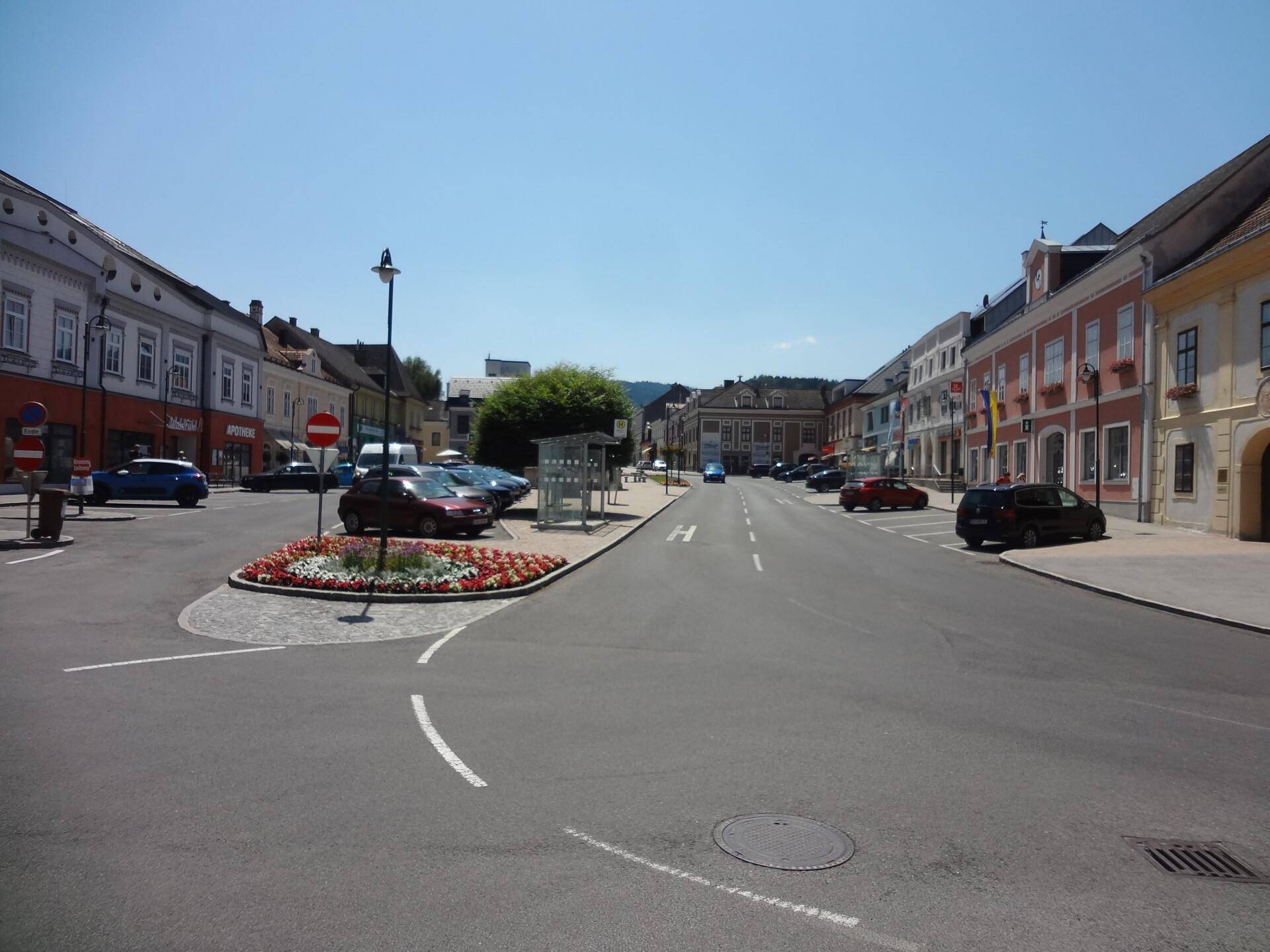 Historischer Stadtplatz mit einem Brunnen, umgeben von Gebäuden und Geschäften, belebt von parkenden Autos.