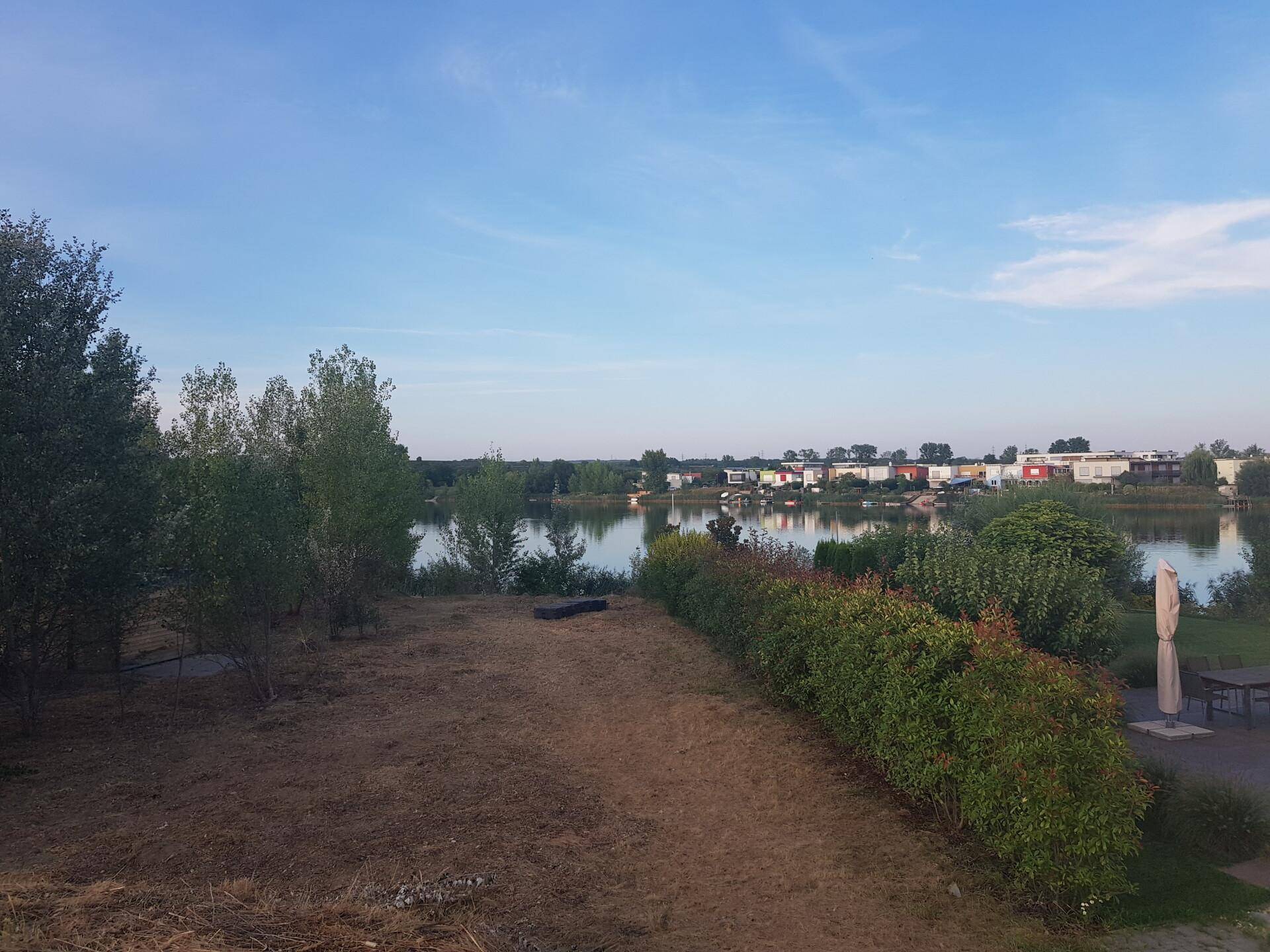 Weitläufiger Blick über einen See mit Siedlung am Ufer und grüner Landschaft.