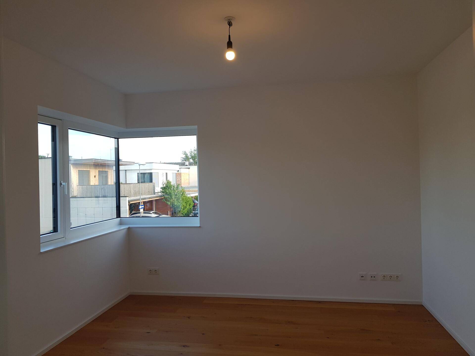 Geräumiges Zimmer mit hellen Wänden, Parkettboden und großen Eckfenstern.