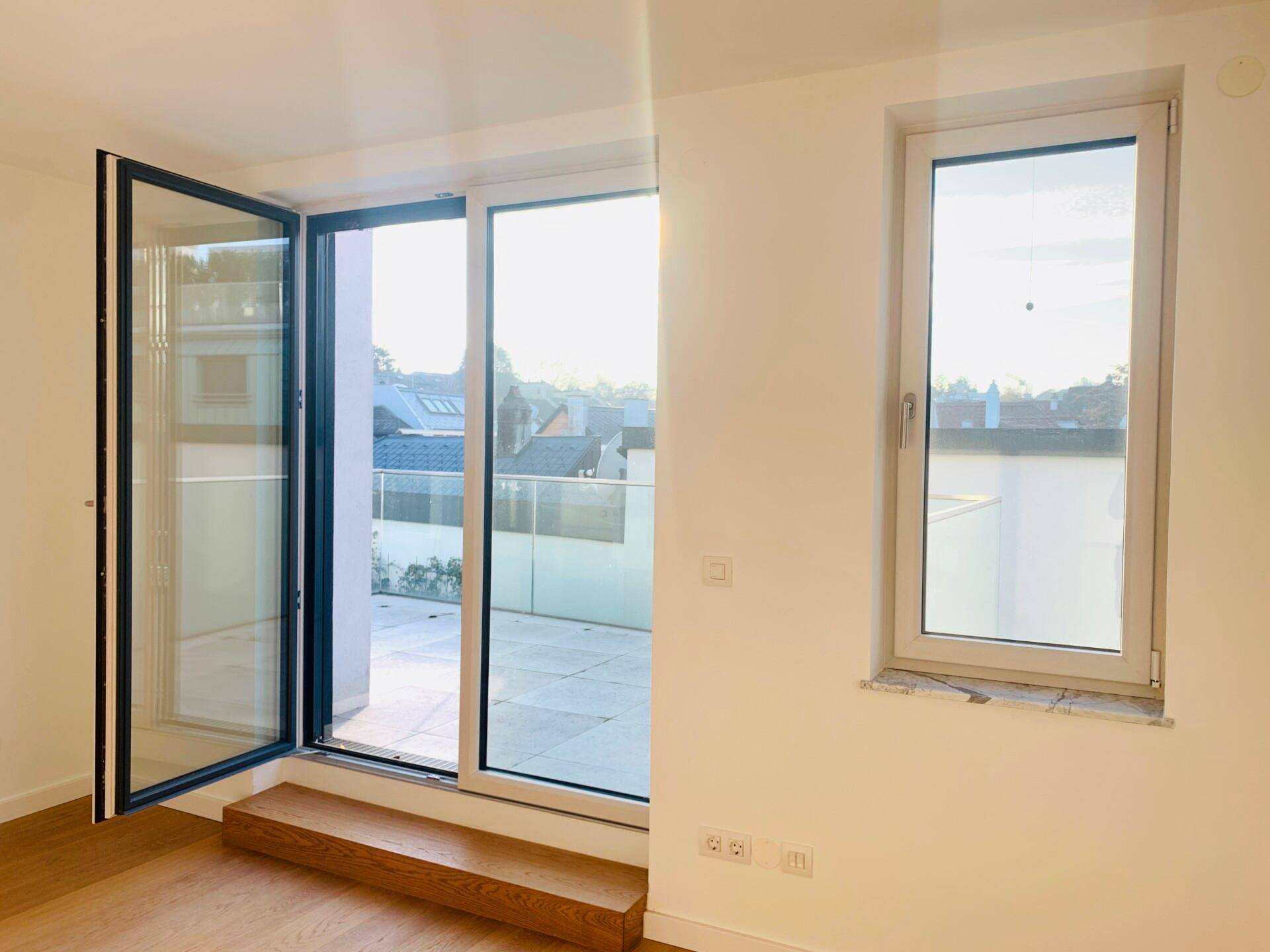 Heller Raum mit Holzboden und großen Fenstern, die Zugang zu einem Außenbereich bieten.