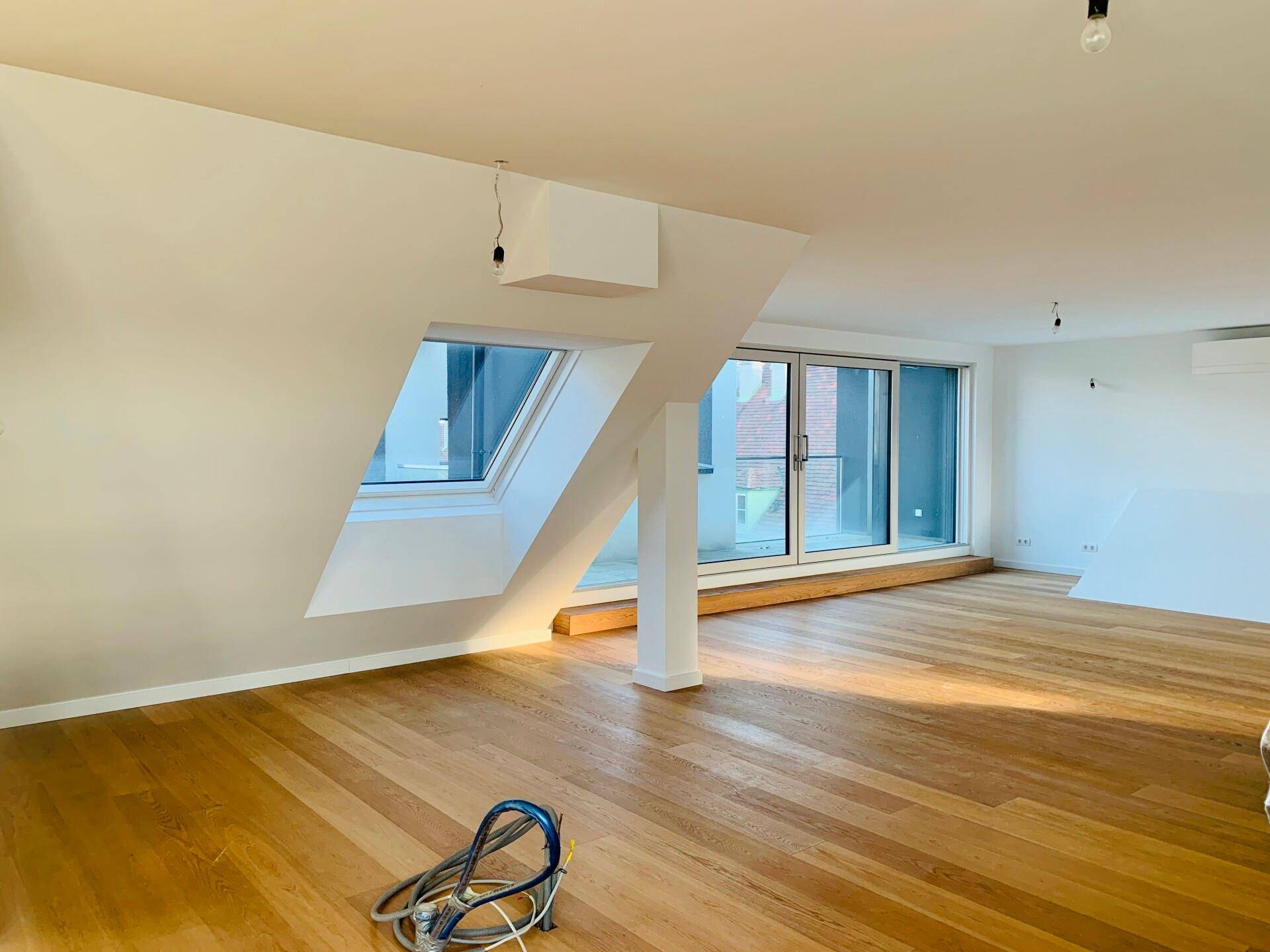 Geräumiger Raum mit Holzboden, Dachschrägen und großen Fenstern, die viel Licht hereinlassen.