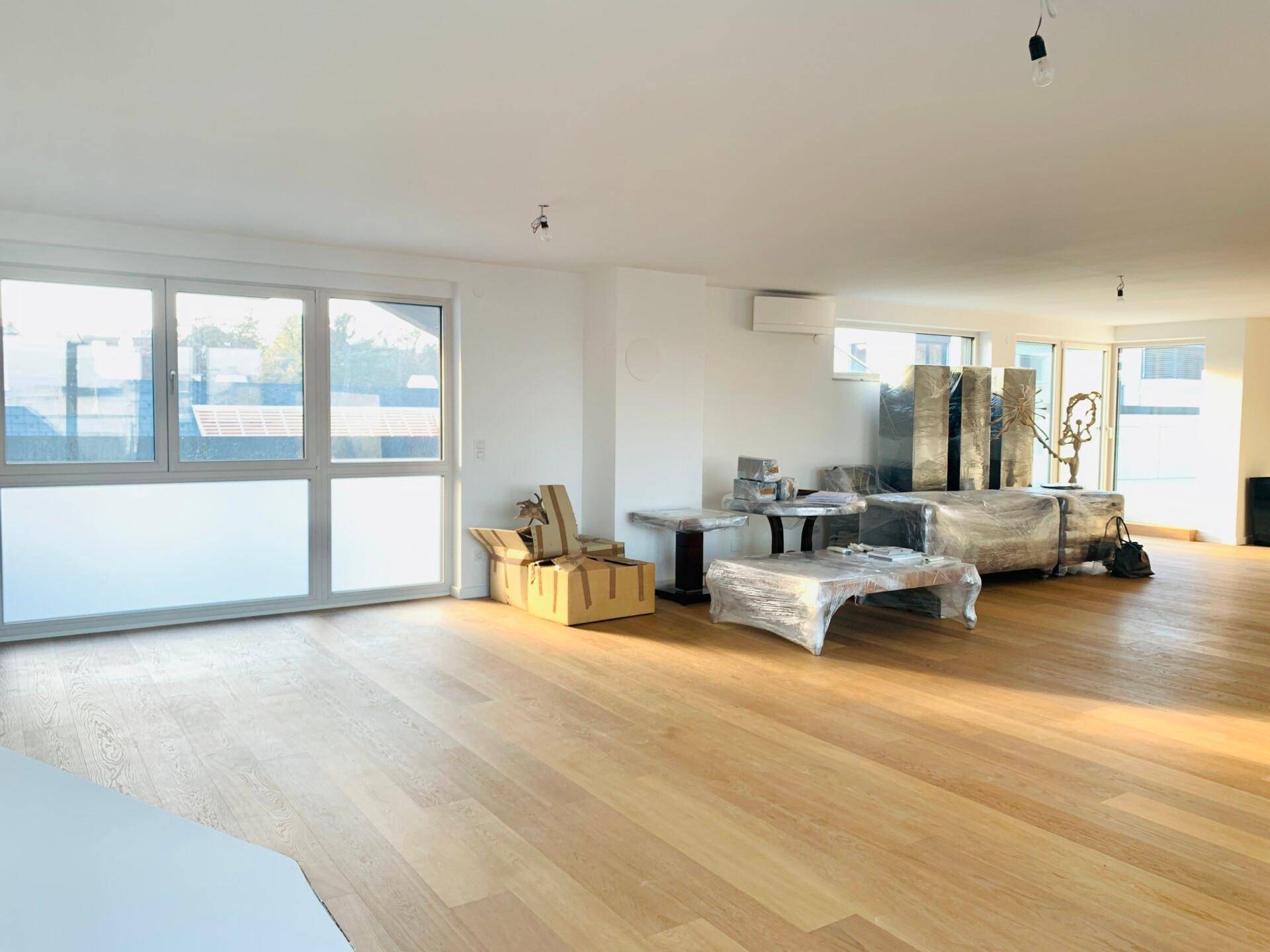 Lichtdurchfluteter Raum mit Holzboden und großen Fenstern, teilweise mit Möbeln abgedeckt und Klimaanlage.