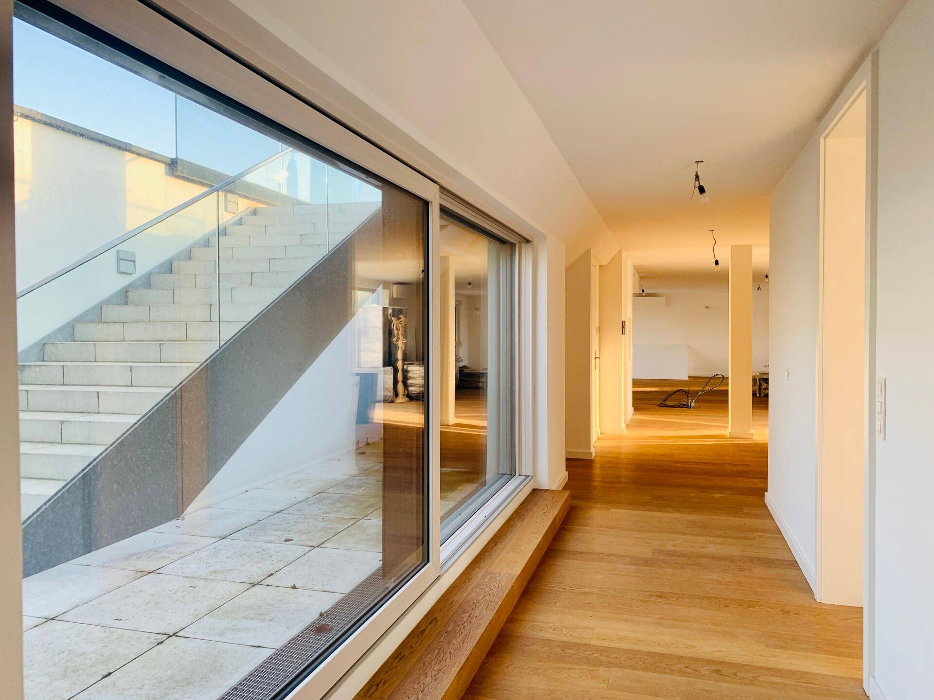 Heller Flur mit Holzboden und großen Fenstern, die eine Treppe und einen Außenbereich zeigen.