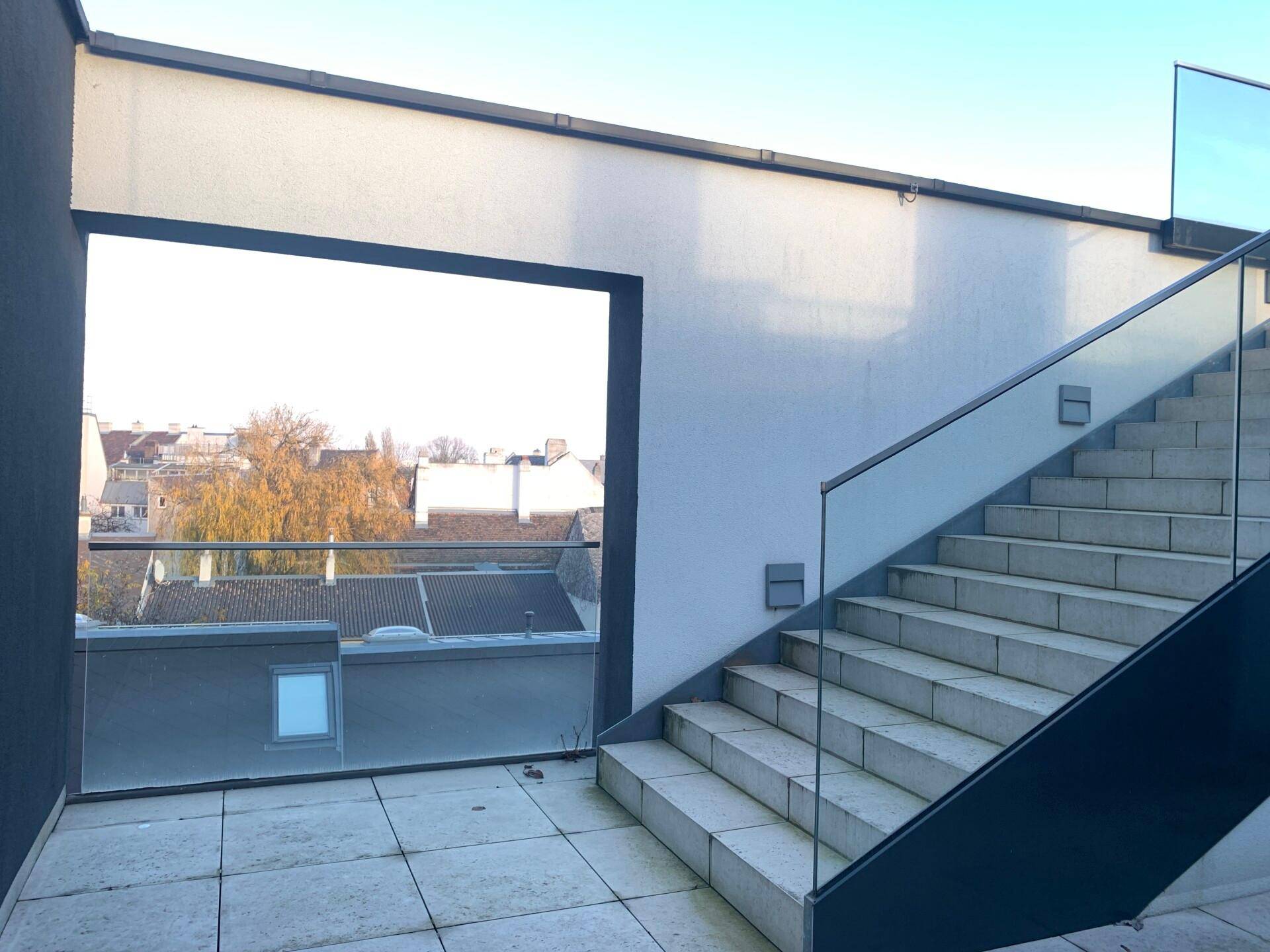 Außentreppe mit Glasgeländer und Blick auf die Dächer der Umgebung unter blauem Himmel.