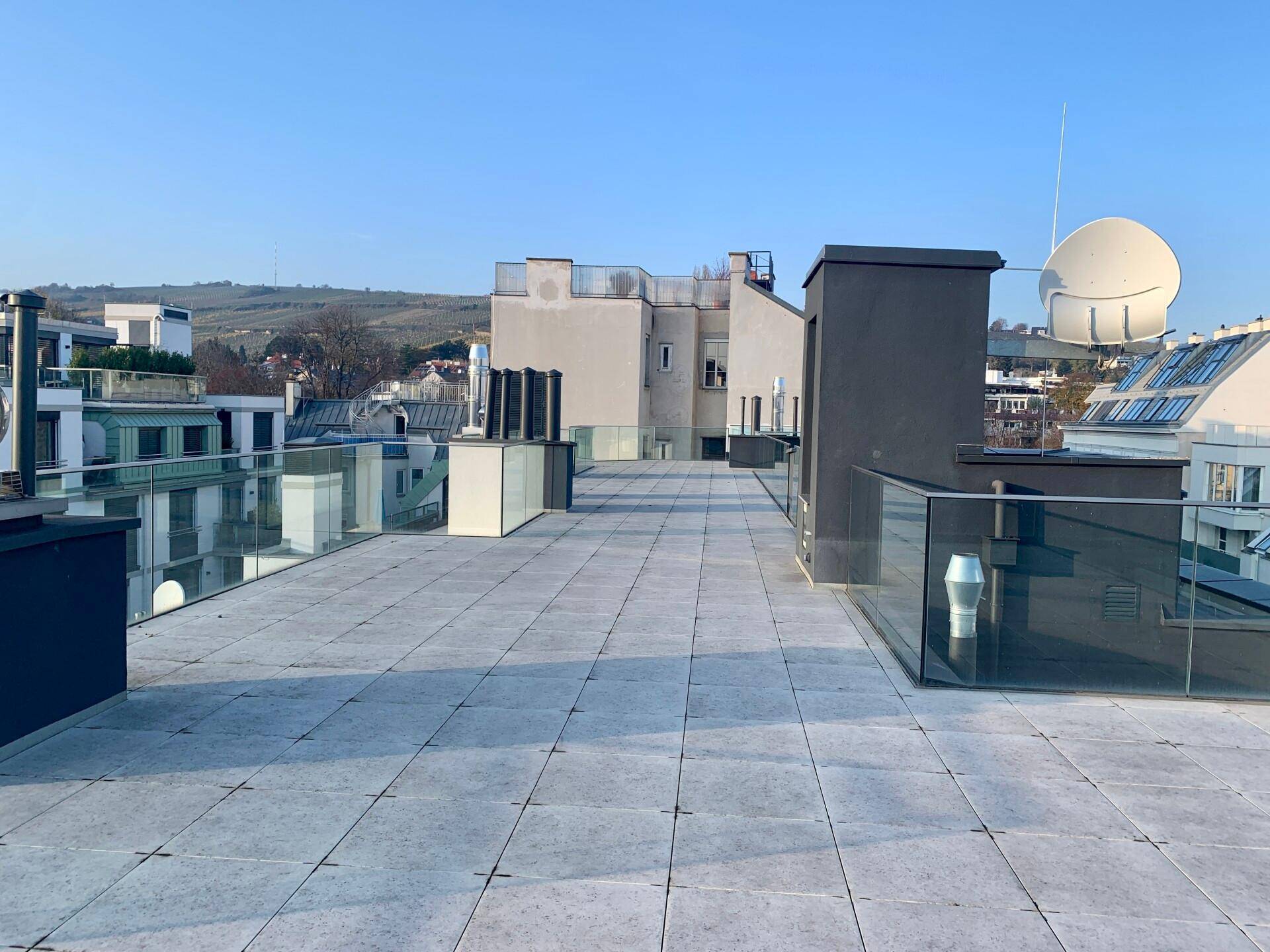 Helle Dachterrasse mit Fliesenboden und Glasgeländer, umgeben von modernen Gebäuden unter blauem Himmel.