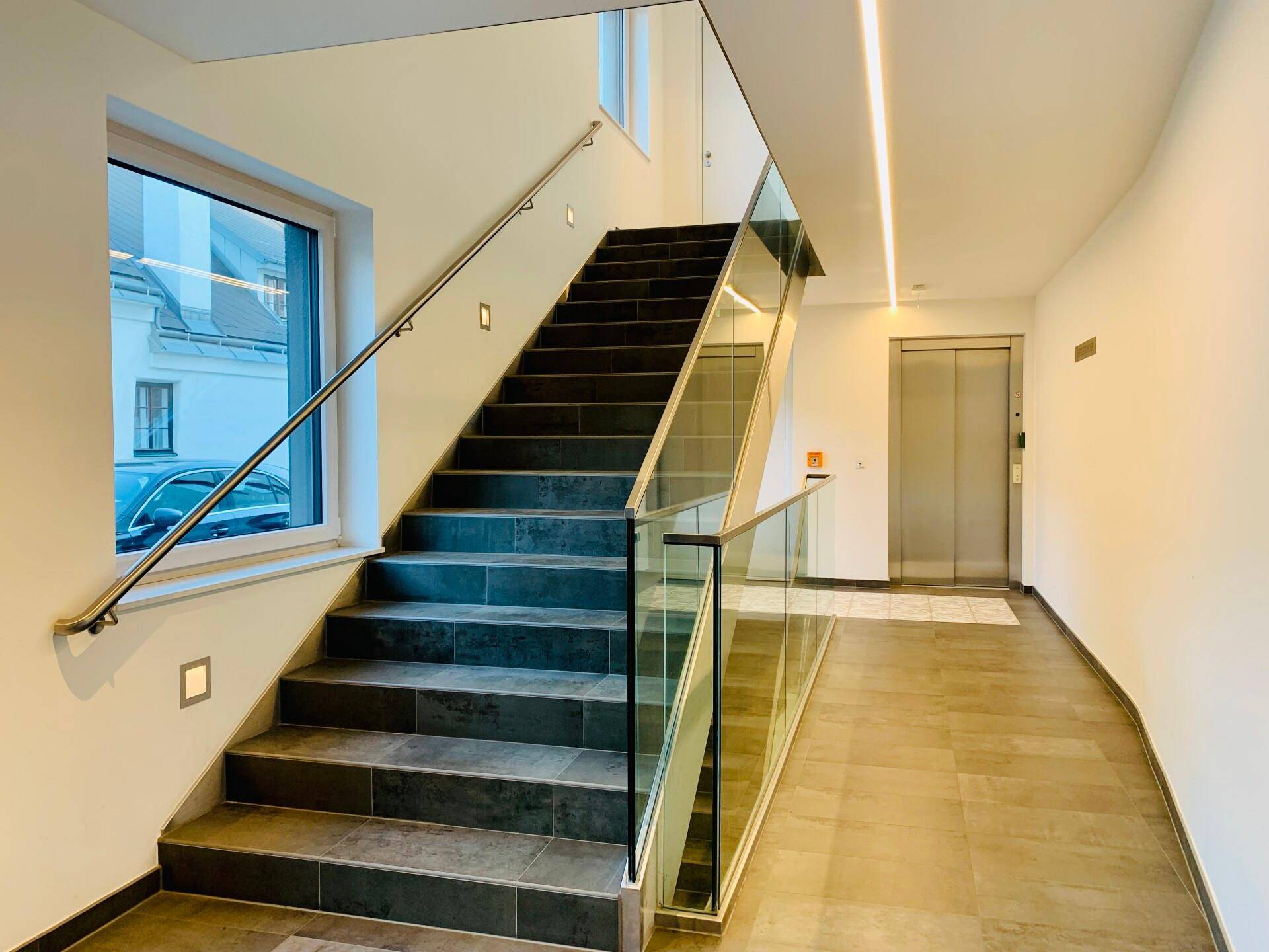 Moderner Eingangsbereich mit Treppe, Glasgeländer und Aufzug, hell beleuchtet.