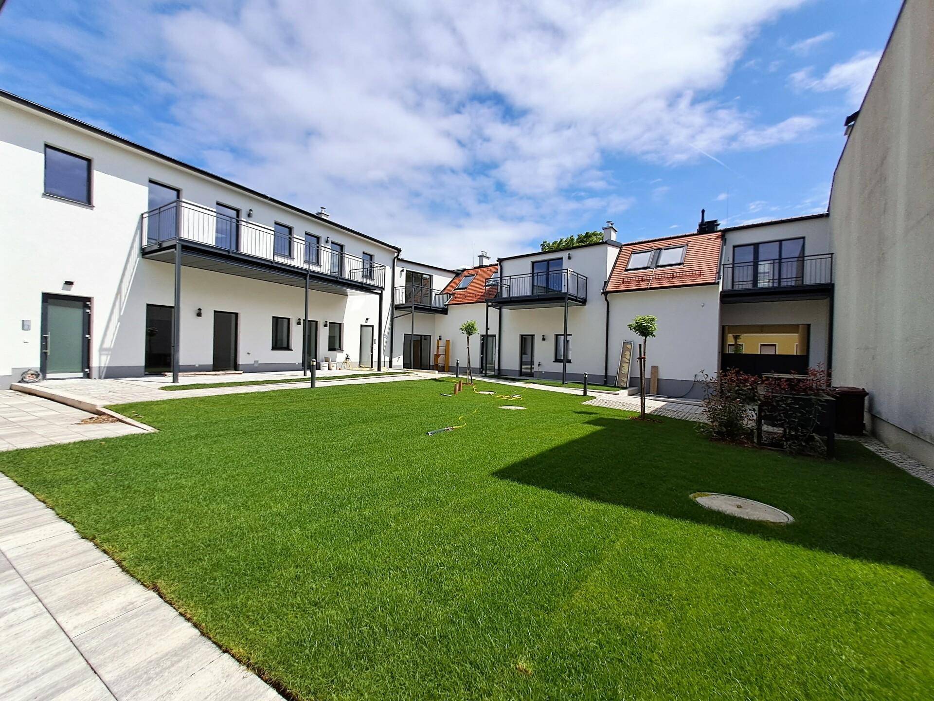 Sonniger Innenhof mit Rasenfläche und modernen Gebäuden mit Balkonen und roten Dächern.