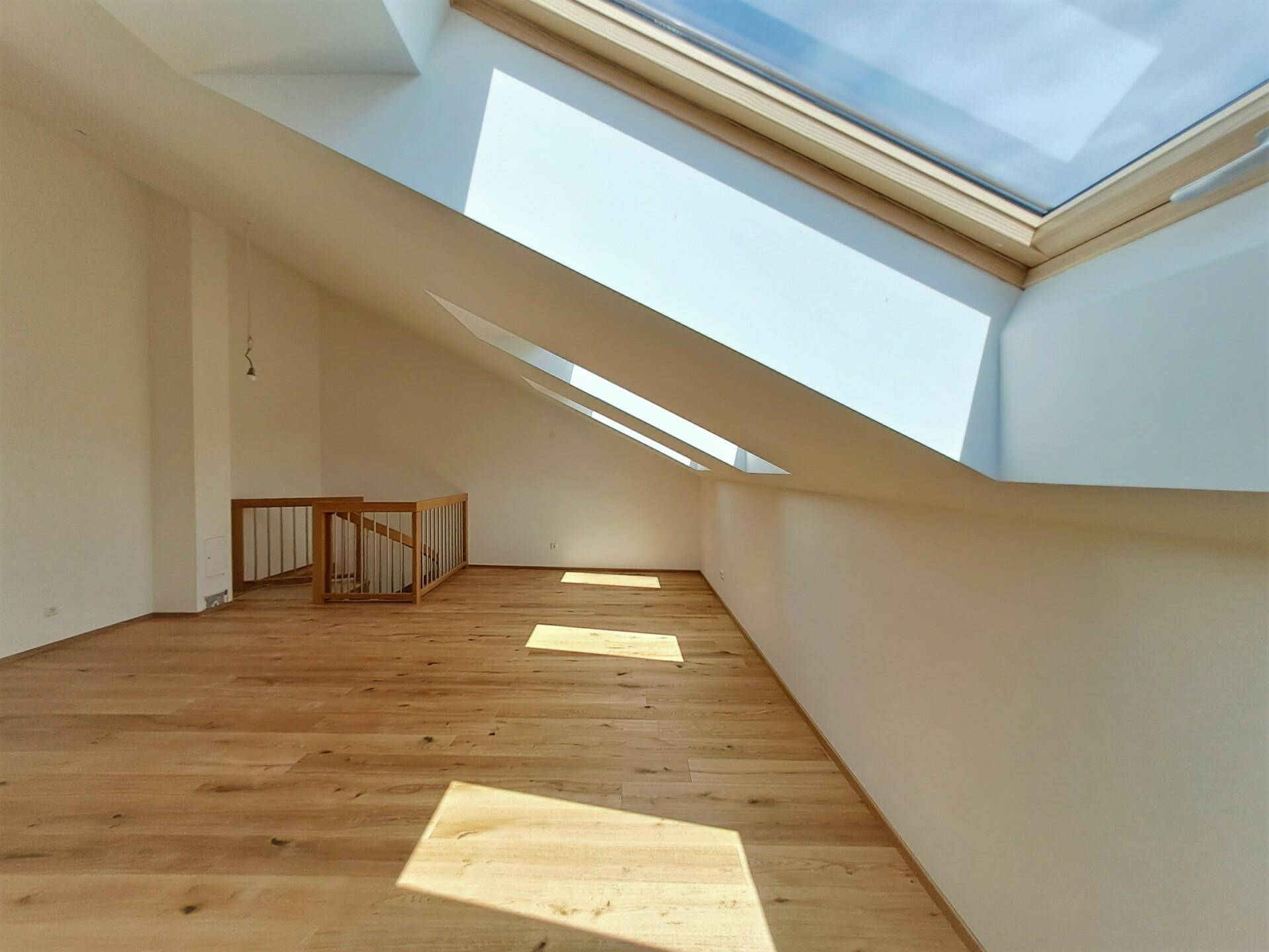 Heller Dachraum mit Holzboden, schrägen Wänden und großen Dachfenstern für viel Tageslicht.
