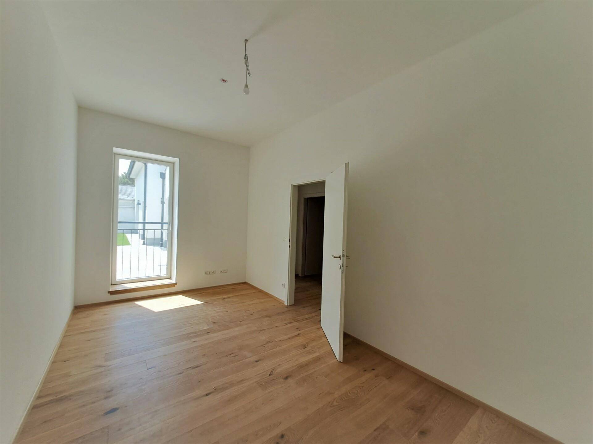 Heller, leerer Raum mit Holzboden und einem Fenster mit Zugang zu einem kleinen Balkon.