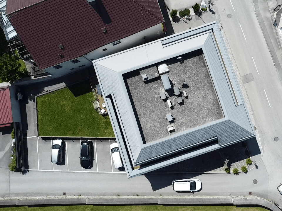 Vogelperspektive eines Gebäudes mit Dachterrasse, Parkplätzen und angrenzendem Gartenbereich.