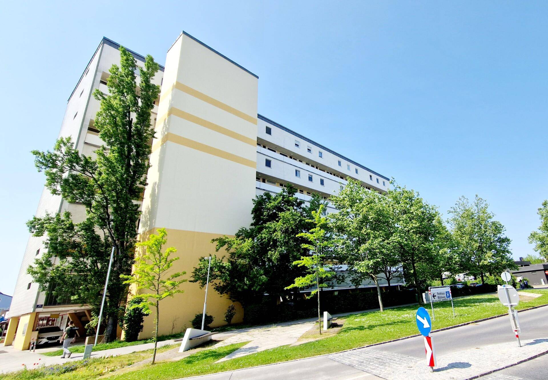 Mehrstöckiges Wohngebäude mit gelben Akzenten und umgebenden Grünflächen unter blauem Himmel.