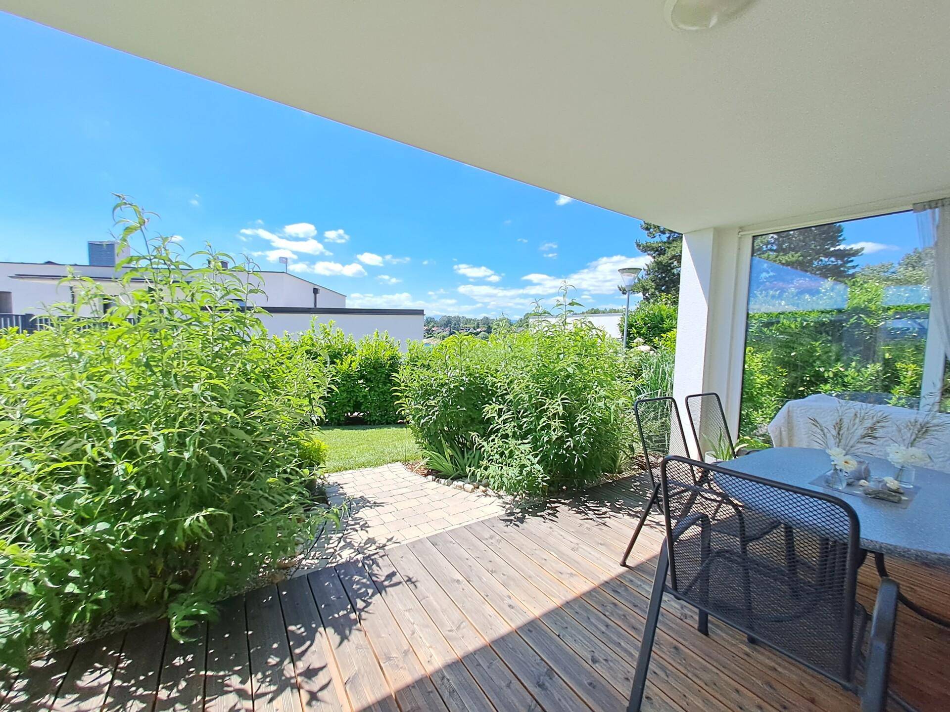 Sonnige Holzterrasse mit Gartenmöbeln, umgeben von üppigen grünen Pflanzen und Sträuchern.