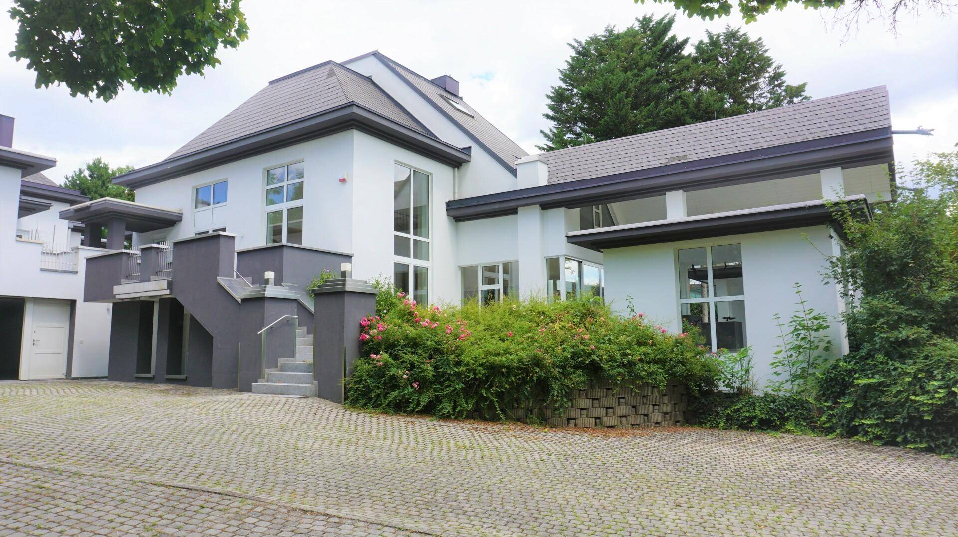 Elegante Villa mit weißer Fassade, grauen Akzenten und gepflasterter Auffahrt umgeben von Grün.