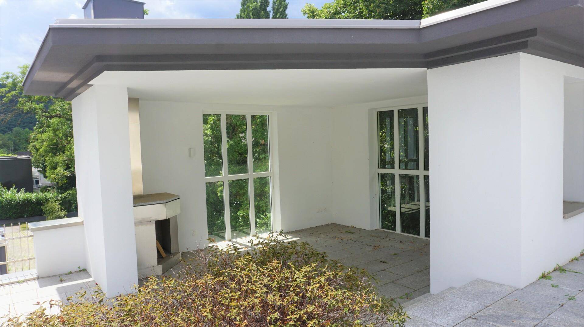 Überdachte Terrasse mit Außenkamin und großen Fenstern, umgeben von üppigem Grün.
