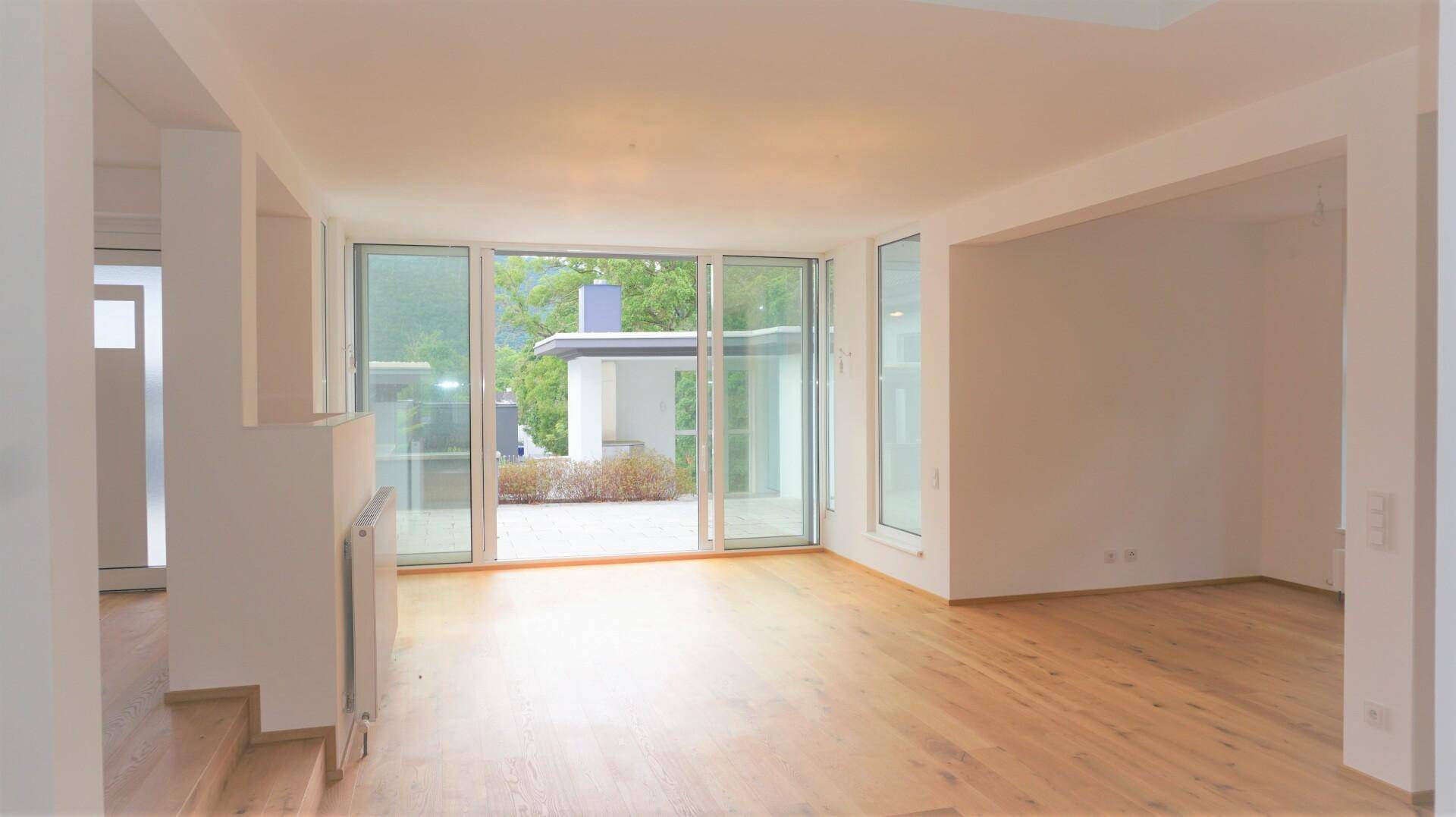 Geräumiger, heller Wohnbereich mit Holzboden und großen Fensterfronten, die viel Licht hereinlassen.