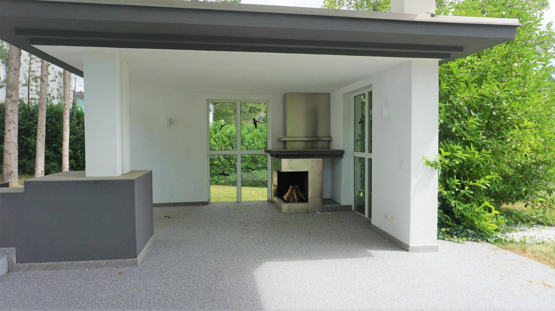 Überdachte Terrasse mit integriertem Grillbereich und Kamin, umgeben von Bäumen.