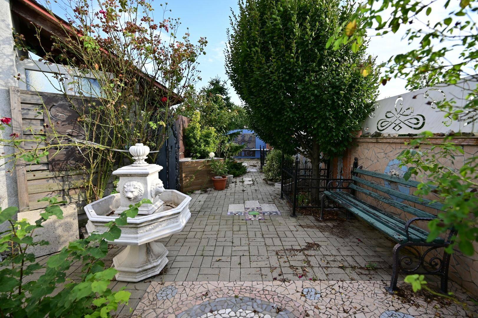 Gepflasterter Gartenweg mit einem dekorativen Brunnen und einer Sitzbank, umgeben von Pflanzen und Bäumen, die zum Verweilen einladen.