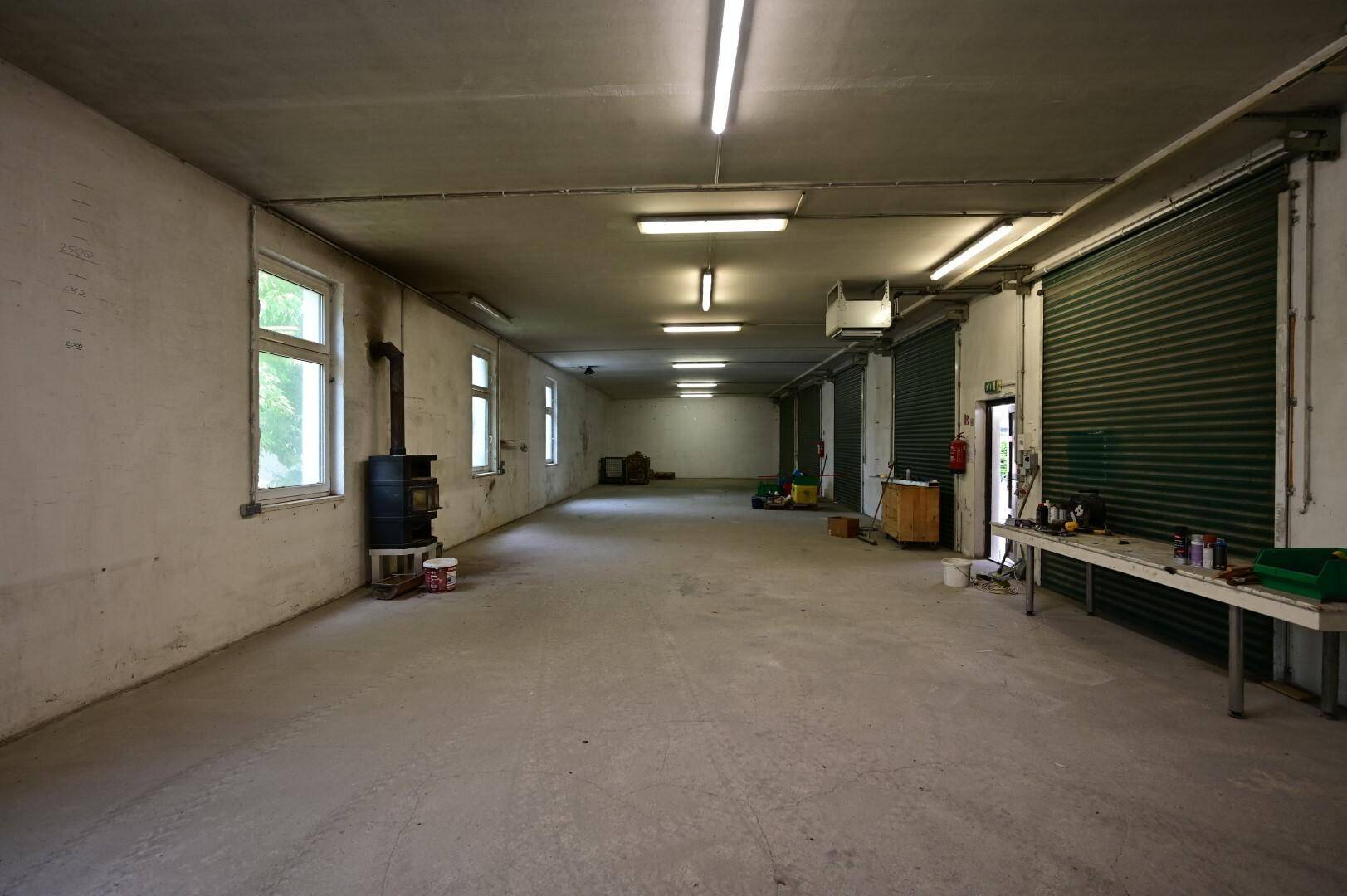 Großer Werkstatt- oder Lagerraum mit Betonboden, Rolltoren und einem Holzofen für Heizung, ideal für handwerkliche Tätigkeiten.