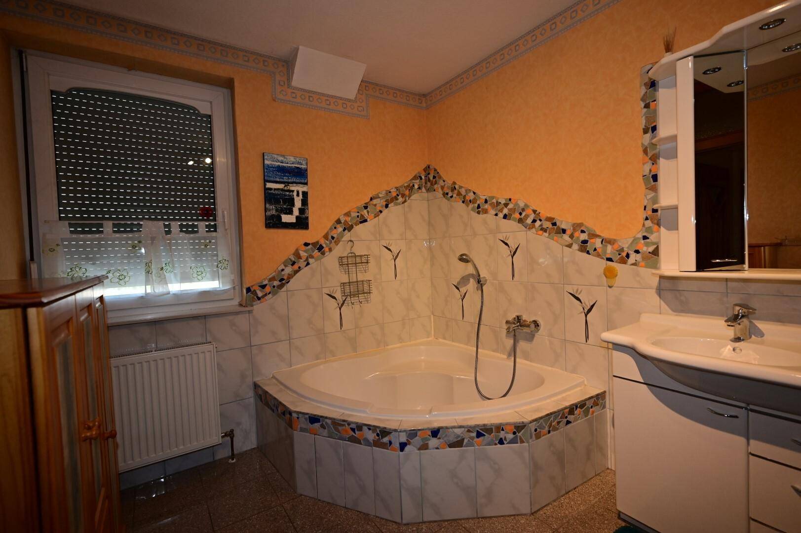 Badezimmer mit Eckbadewanne, Fliesenboden und einem Fenster, das Tageslicht hereinlässt und für Belüftung sorgt.