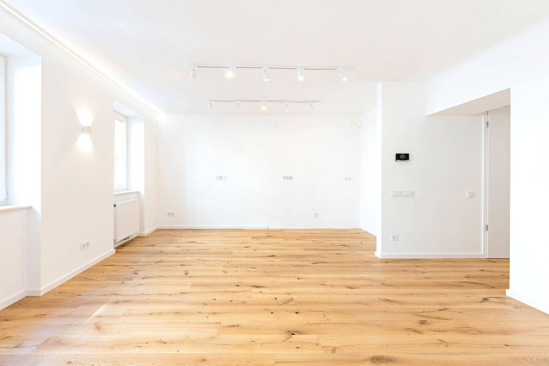 Geräumiger, leerer Raum mit hellem Holzboden, großen Fenstern und moderner Deckenbeleuchtung.