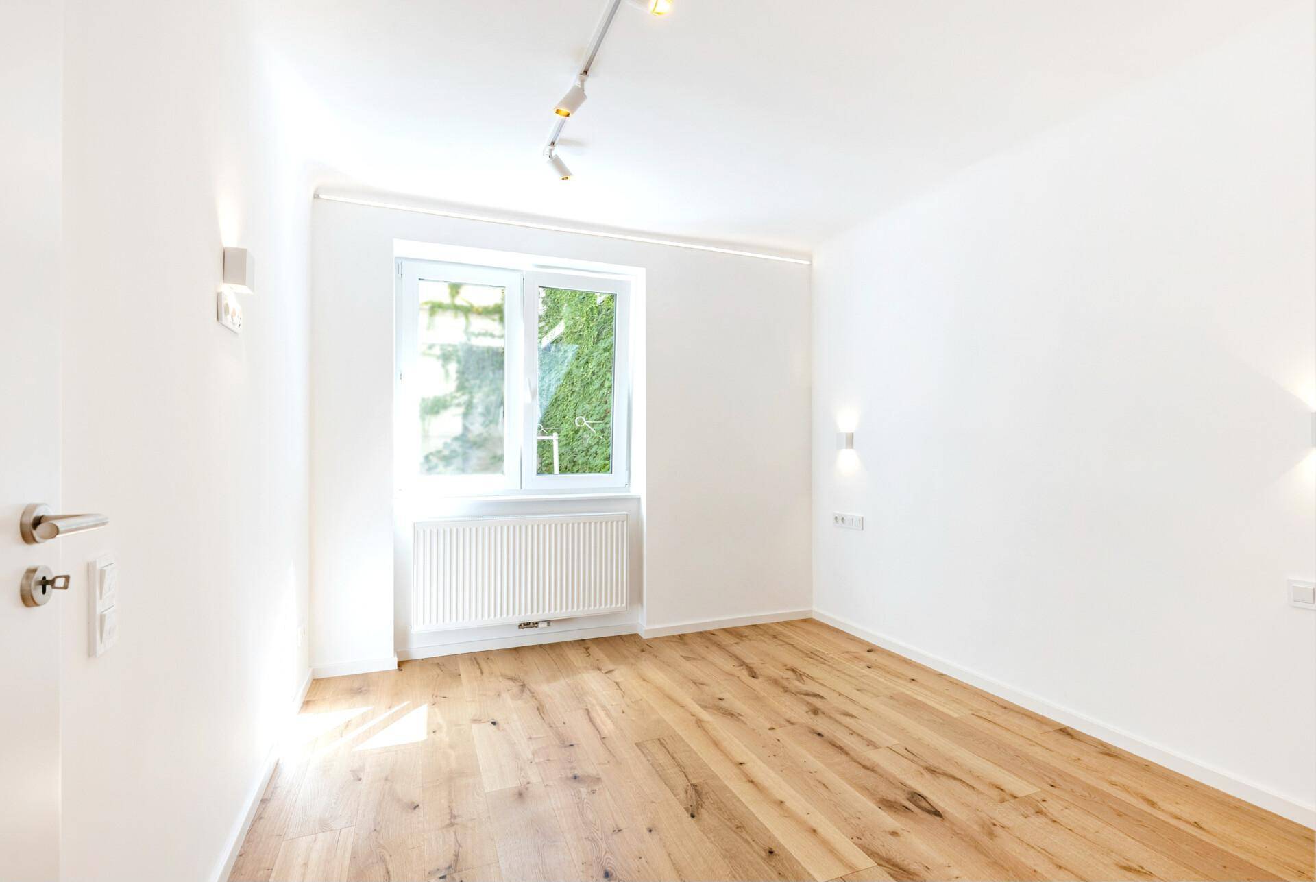 Helles Zimmer mit Holzboden, einem Fenster mit Blick ins Grüne und weiß gestrichenen Wänden.