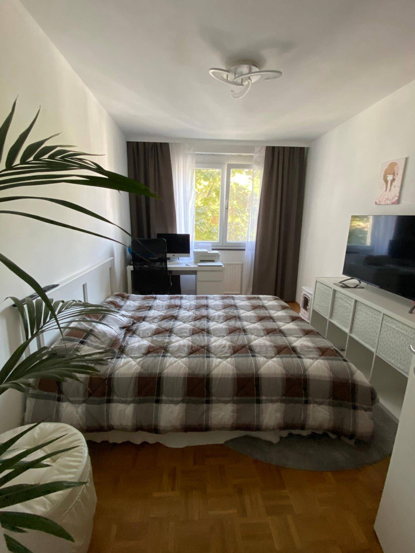 Helles Schlafzimmer mit Parkettboden, großem Bett, Schreibtisch am Fenster und einer Zimmerpflanze.