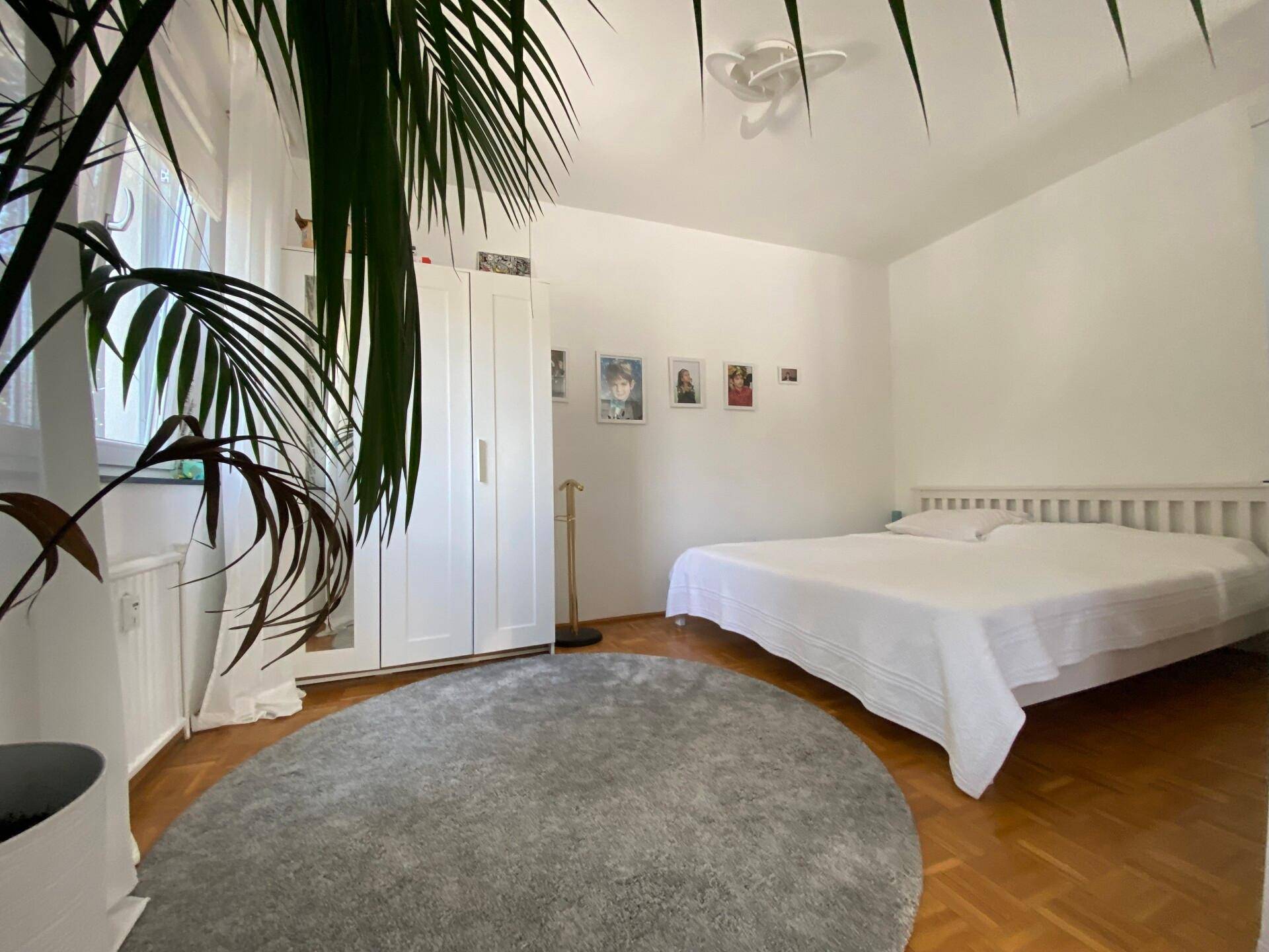 Helles Schlafzimmer mit Parkettboden, weißem Bett und Kleiderschrank, dekoriert mit einer großen Zimmerpflanze.