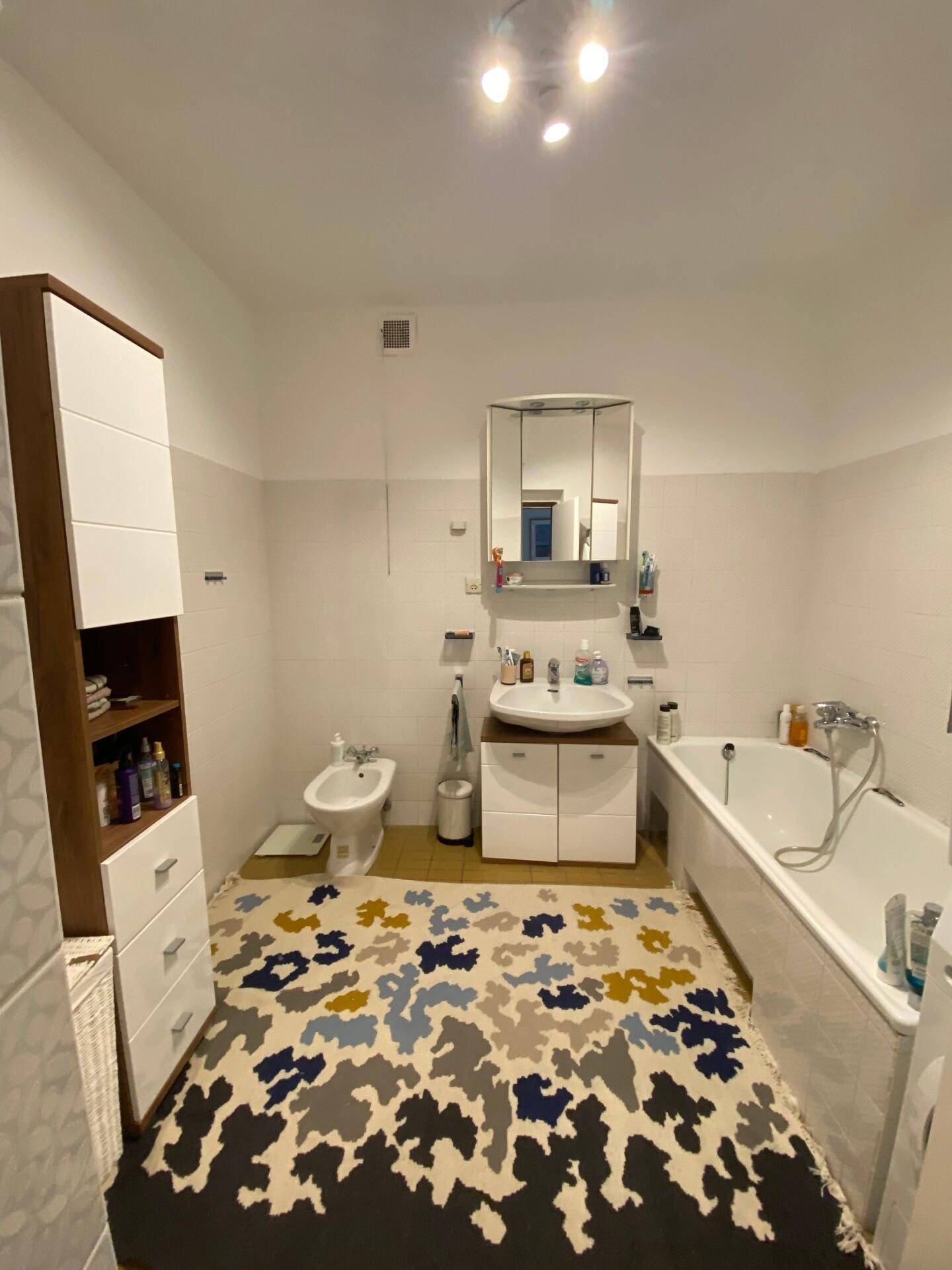 Badezimmer mit Badewanne, Bidet, Waschbecken und einem farbenfrohen Teppich auf dem Fliesenboden.
