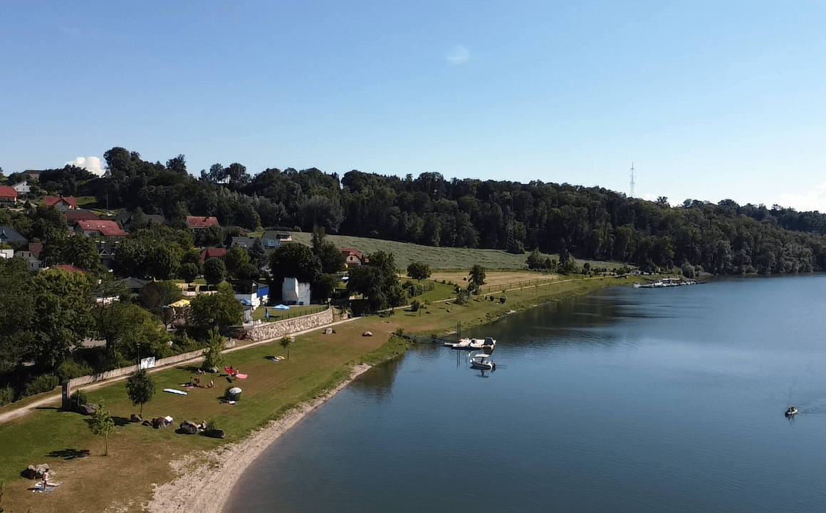 Malerische Uferlandschaft mit Badestrand, Häusern und dicht bewaldeten Hügeln im Hintergrund.