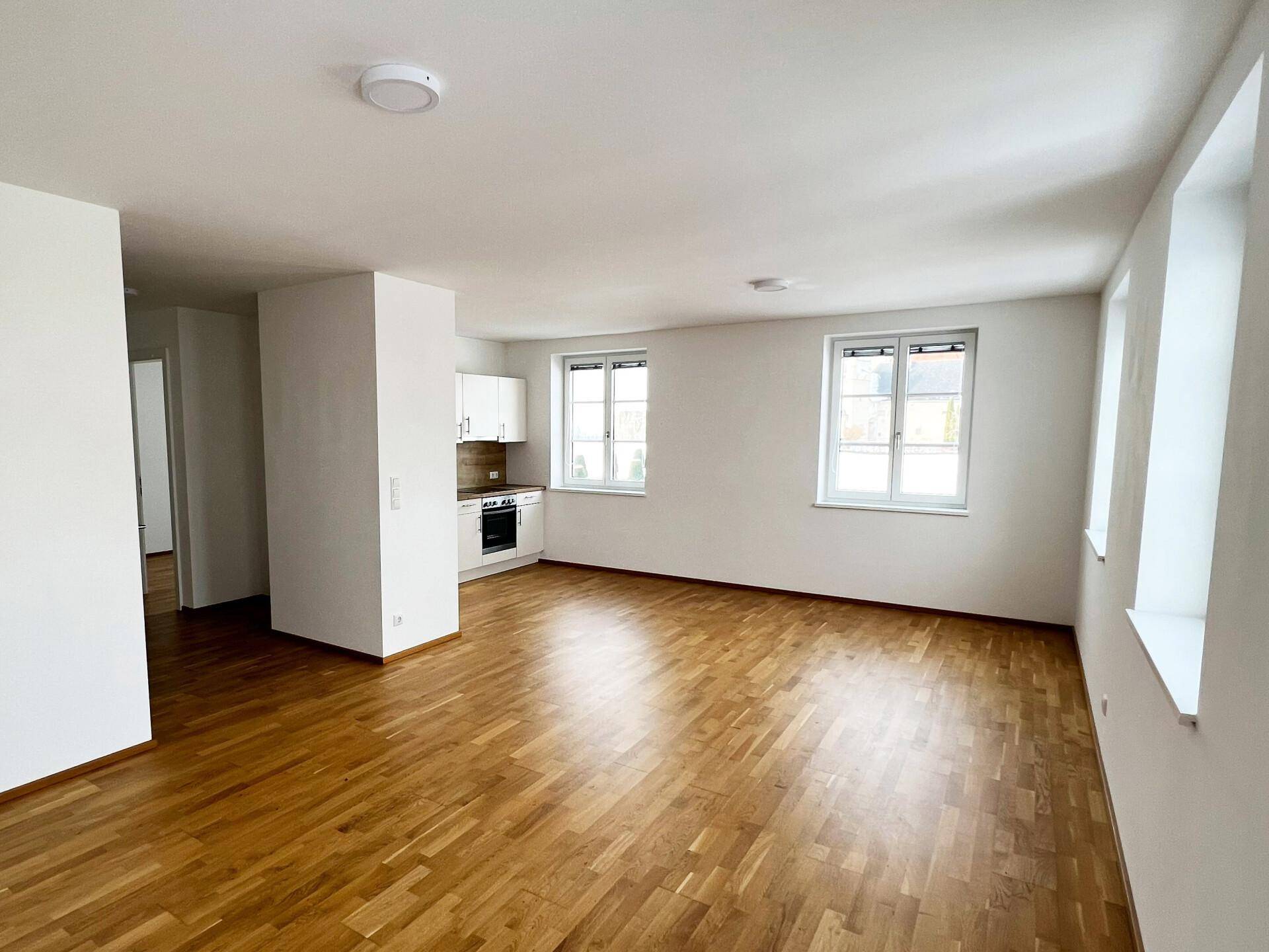 Geräumiger Wohnbereich mit Holzboden, großen Fenstern und integrierter Küchenzeile.